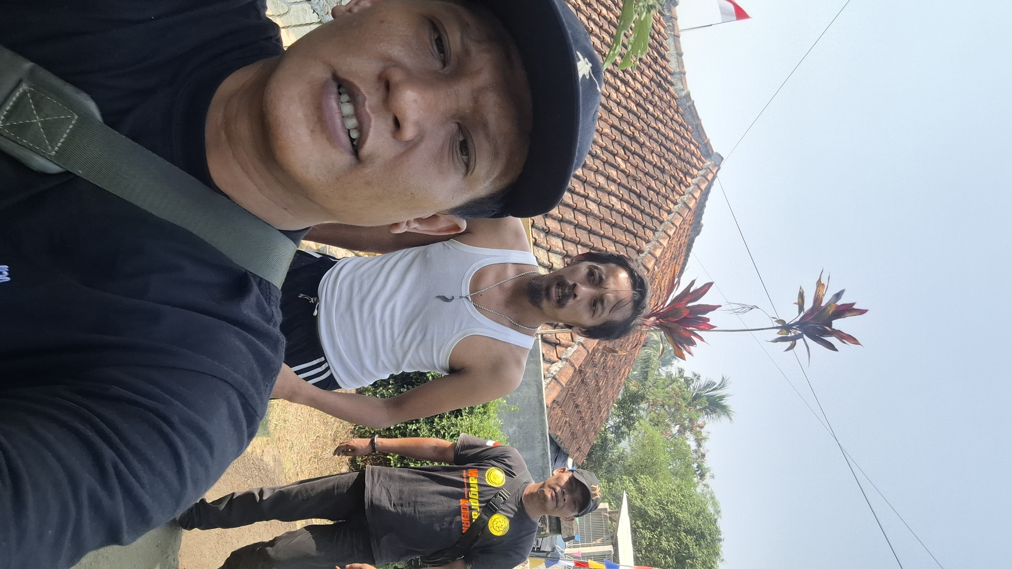 In Rahong, Indonesia, a selfie is taken outdoors during the day. Three men are visible, with the closest one in the foreground wearing a black t-shirt and a black baseball cap. The second man, positioned behind and to the right of the first, is wearing a white tank top and black shorts with white stripes. The third man, to the right and further back, is wearing a black t-shirt and dark pants with a black baseball cap. Behind the men, a red-tiled roof of a building is prominent, with an Indonesian flag visible on the left side. Tropical plants with red and green leaves are present, along with trees and a blue sky. The ground appears to be a dirt path.