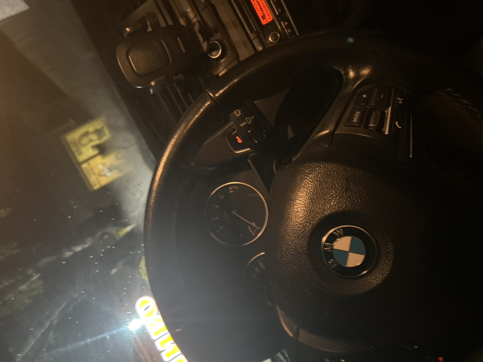 The image shows the interior of a car, viewed from the driver's perspective. The steering wheel, dashboard, and part of the center console are visible. The BMW logo is prominently displayed on the steering wheel. On the dashboard, a speedometer and tachometer are visible, as are controls for the windshield wipers and radio. A phone mount is attached to the windshield. The scene appears to be at night, with some lighting from outside the car visible on the windshield. The overall impression is a typical car interior at night, with no unusual or remarkable features.