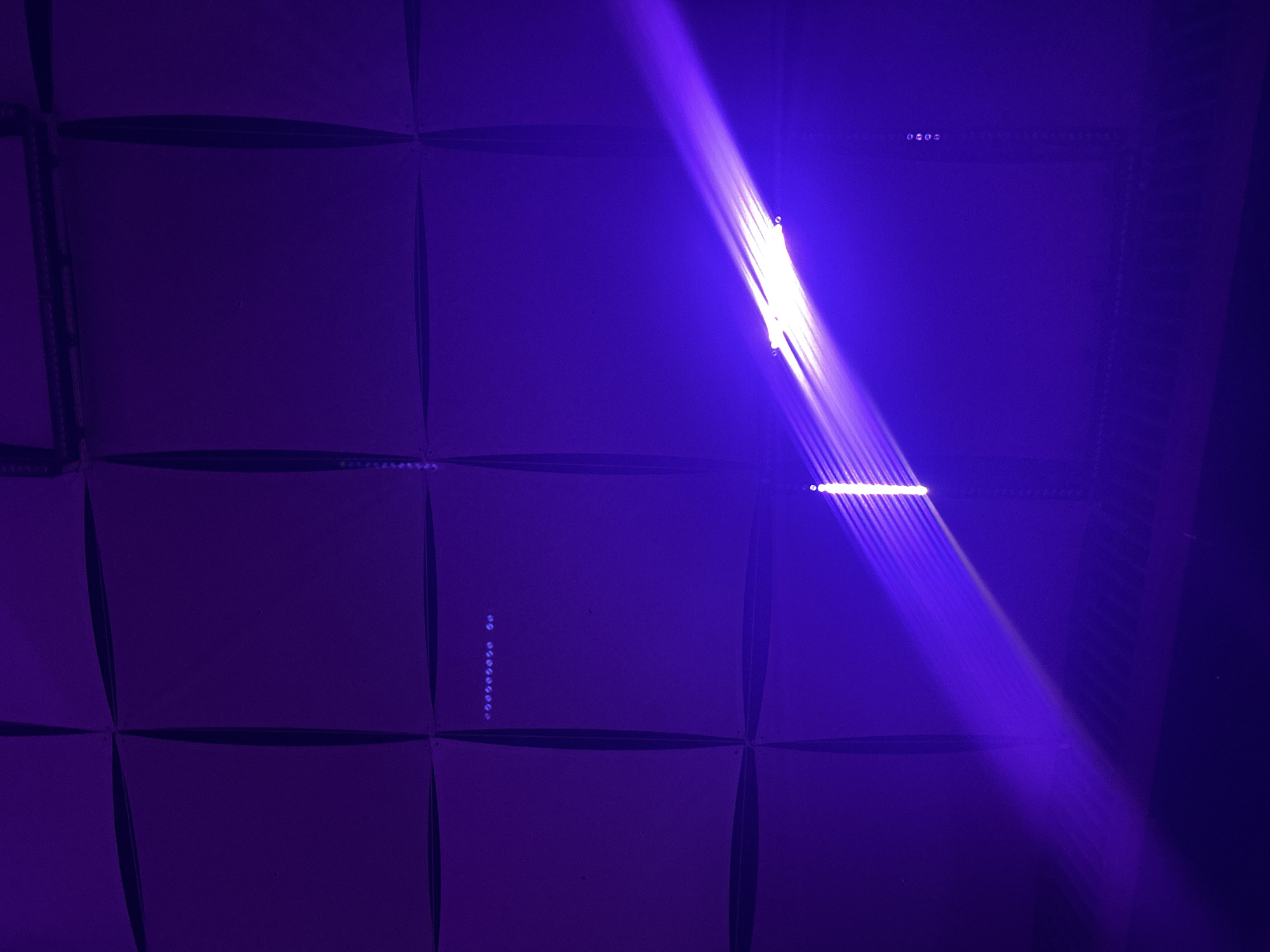 A purple-hued photograph depicts an interior space with a textured, grid-like ceiling or wall. A prominent diagonal beam of bright white and purple light cuts across the scene from the bottom left to the upper right, creating a lens flare effect and illuminating a section of the surface. The surface itself is divided into square panels, some of which have faint, linear patterns or rows of small dots visible within them. These dots appear to be small lights or markers. The overall atmosphere is dark and possibly a venue or stage area, given the dramatic lighting. The image does not contain any discernable people or specific identifiable landmarks that would allow for pinpointing a location within Burgas, Bulgaria.