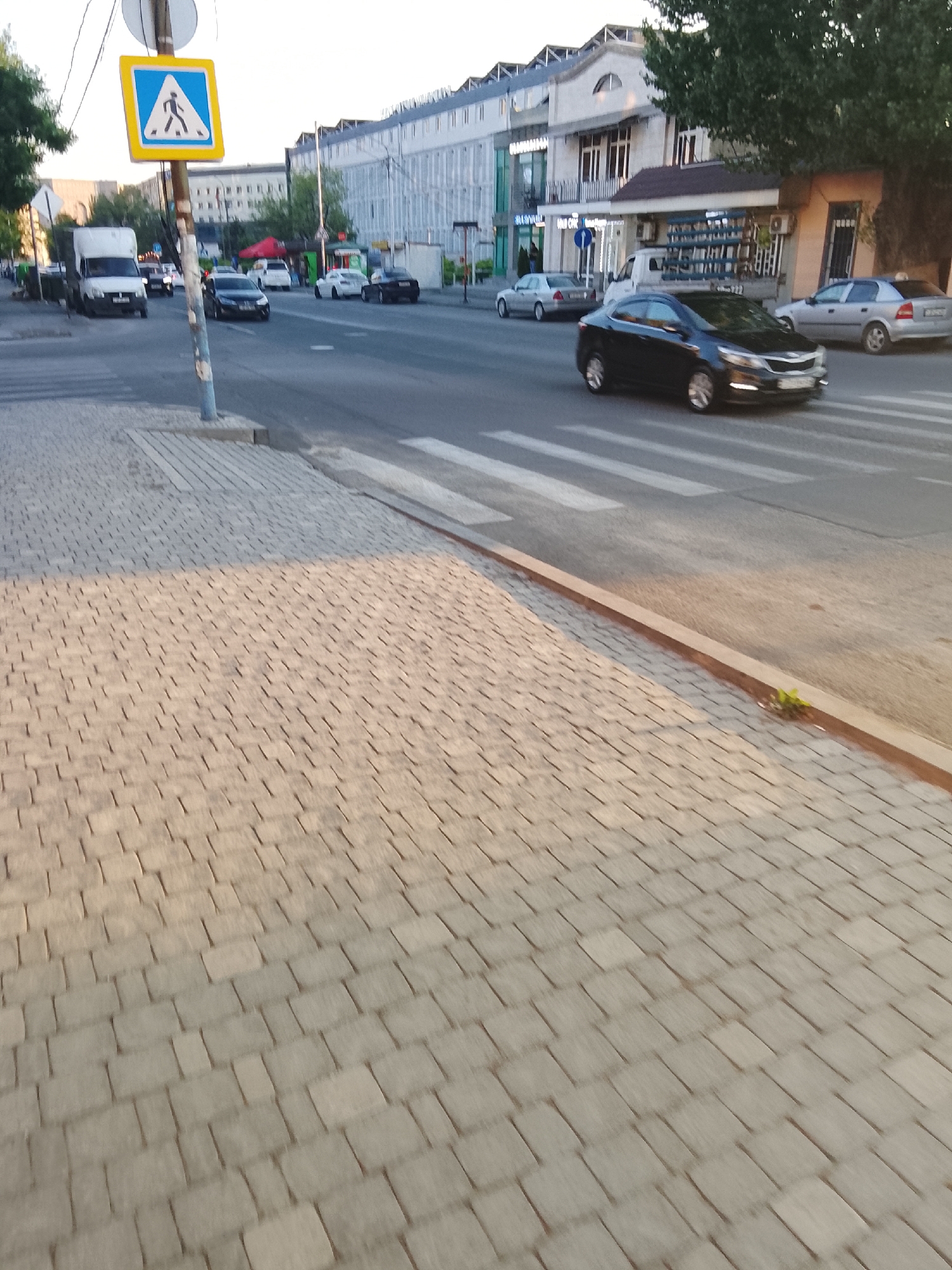 The image depicts a street scene at what appears to be late afternoon or early evening, given the warm lighting and long shadows. The foreground is dominated by a textured cobblestone sidewalk, with a section of lighter-colored pavers in the upper left. A pedestrian crossing sign, featuring a blue square with a white border and a white silhouette of a person walking, is prominently displayed on a pole on the left side of the image.
The street itself is paved and shows clear white lines for a pedestrian crossing. Several cars are visible on the road. A white van is parked on the left, and a black sedan is in motion on the right side of the street, crossing the pedestrian area. Other cars, including a silver sedan, are further down the road.
Behind the street, buildings line the roadside. The architecture varies, with some structures appearing to be commercial establishments with awnings and large windows, while others are more residential in appearance. Trees with full green foliage are also present, contributing to the urban park-like setting. One particularly large tree shades a portion of the sidewalk in the upper right.
The scene suggests a moderately busy urban environment. There are no people clearly visible in the image, though the presence of the pedestrian crossing sign implies their potential presence. The overall mood is calm and ordinary, characteristic of a typical day in a city. No specific text is legible in the image.