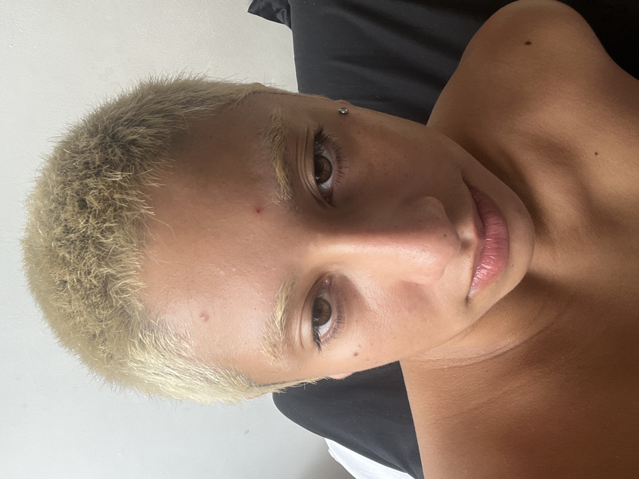 The image presents a close-up, frontal portrait of an individual from the chest upwards. The person has a tan complexion, short-cropped, light blonde or bleached hair, and dark brown eyes. Their eyebrows are light and sparse. Two small red blemishes are visible on the forehead. A silver stud piercing is present on the left temple, lateral to the left eye. The individual's shoulders and upper chest are visible, suggesting bare skin or minimal clothing. The lips are closed and exhibit a natural pinkish hue. The expression is direct, with a slight downward gaze. The background on the left consists of a light, uniform surface, possibly a wall, while the right side shows a dark, indistinct fabric, likely a pillow or bedding. The scene appears to be indoors, possibly in a private residence.
The specific location within the city of Buenos Aires, Argentina, cannot be determined from the visual evidence provided in this close-up indoor portrait. No architectural details, landmarks, or identifying environmental features are present.