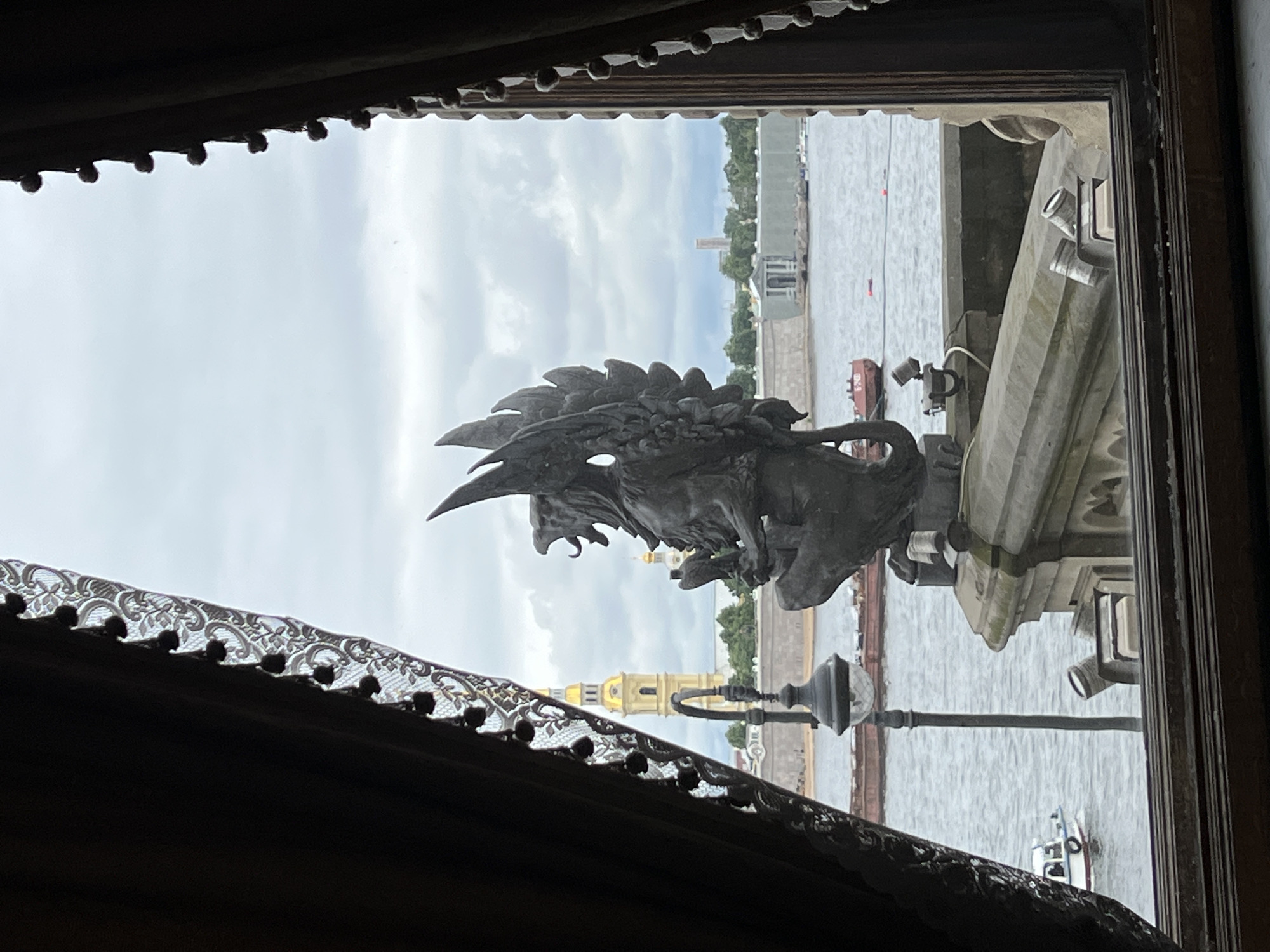 The image is an outdoor, eye-level, full shot taken through a window with heavy curtains framing the view. The scene appears to be St. Petersburg, Russia. In the foreground, a dark, ornate curtain with tassels and lace detailing frames the left and right sides of the view. A stone statue of a winged creature, possibly a griffin, with its mouth open and wings spread, is perched on a stone ledge in the middle of the frame.
Beyond the statue, a body of water, likely a river or canal, is visible. A dark, flat-bottomed barge is moored on the water to the left of the statue, and a small white boat with a blue stripe is moving away from the viewer on the far left. A lamppost with a glass globe is positioned in front of the water.
In the background, a tall, yellow bell tower with a golden dome and spire dominates the skyline on the left side of the frame. Further in the distance, a long, light-colored building with architectural details is visible, and a modern, rectangular tower rises on the right. The sky is overcast with grey clouds. The overall atmosphere is somber and slightly muted due to the weather and the framing of the shot.