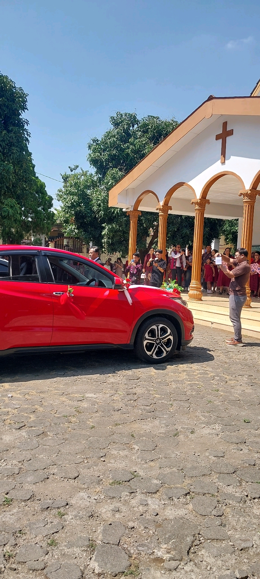 In Keling, Indonesia, a red car is parked on a cobbled street in front of a building with a cross at its peak. The car is decorated with white ribbons. A group of people, some wearing red outfits, stand near the building's entrance, while others take photos. One person in the foreground is holding a phone and appears to be recording. The scene suggests a celebratory event, possibly a wedding or a religious ceremony, taking place outdoors under a clear blue sky.