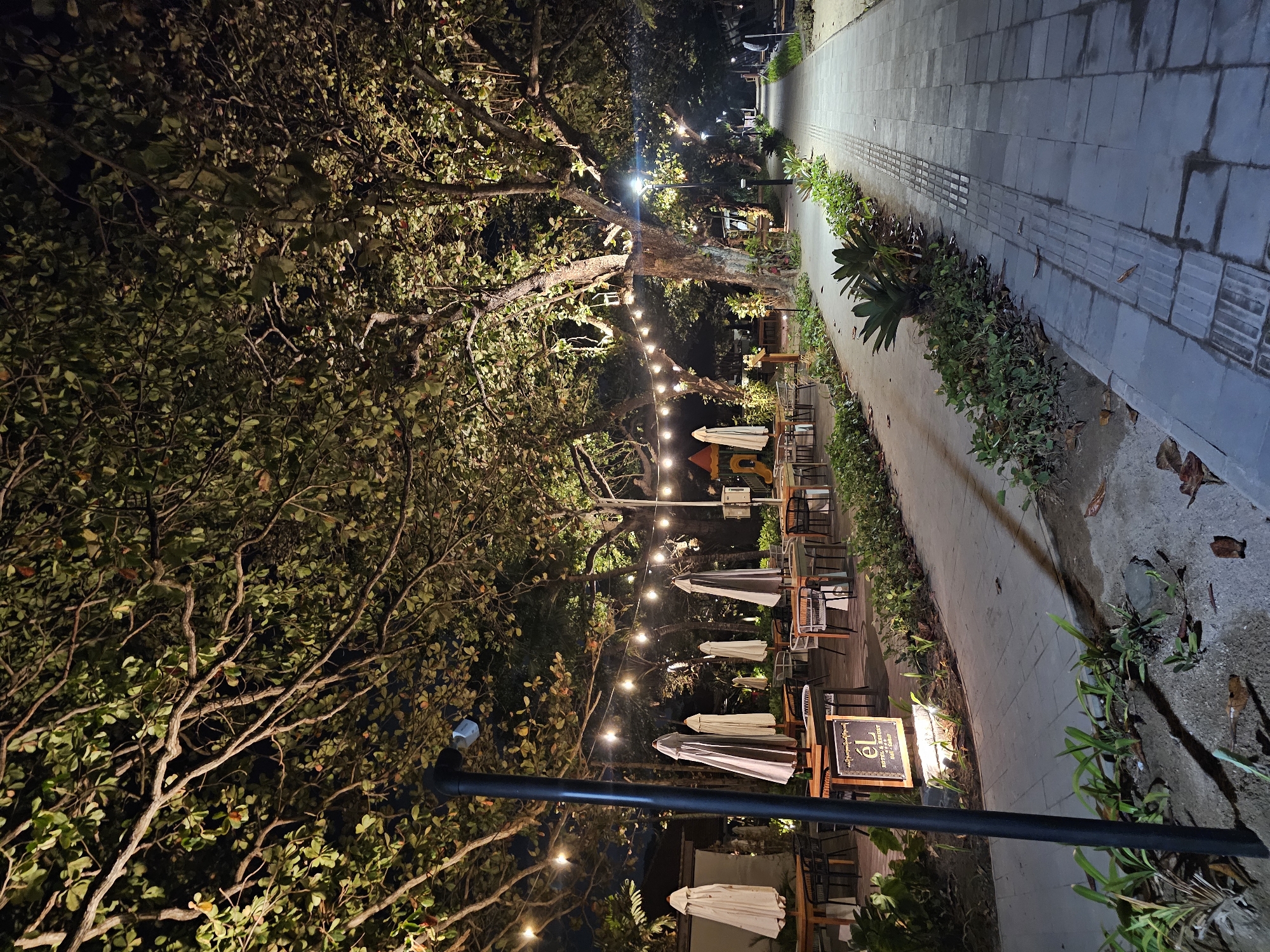 A nighttime view of an outdoor dining area and walkway in Denpasar, Indonesia, is shown. The scene is illuminated by string lights and ground lights, casting a warm glow on the lush foliage and trees overhead. Tables and chairs are neatly arranged, with several closed umbrellas providing shade during the day. A sign for "EL HOTEL & RESORT" is visible near the entrance to the dining area. The walkway, paved with stone tiles, leads into the distance, lined with manicured plants and a textured strip for accessibility. No people are actively engaged in activities, and the environment appears serene and well-maintained.