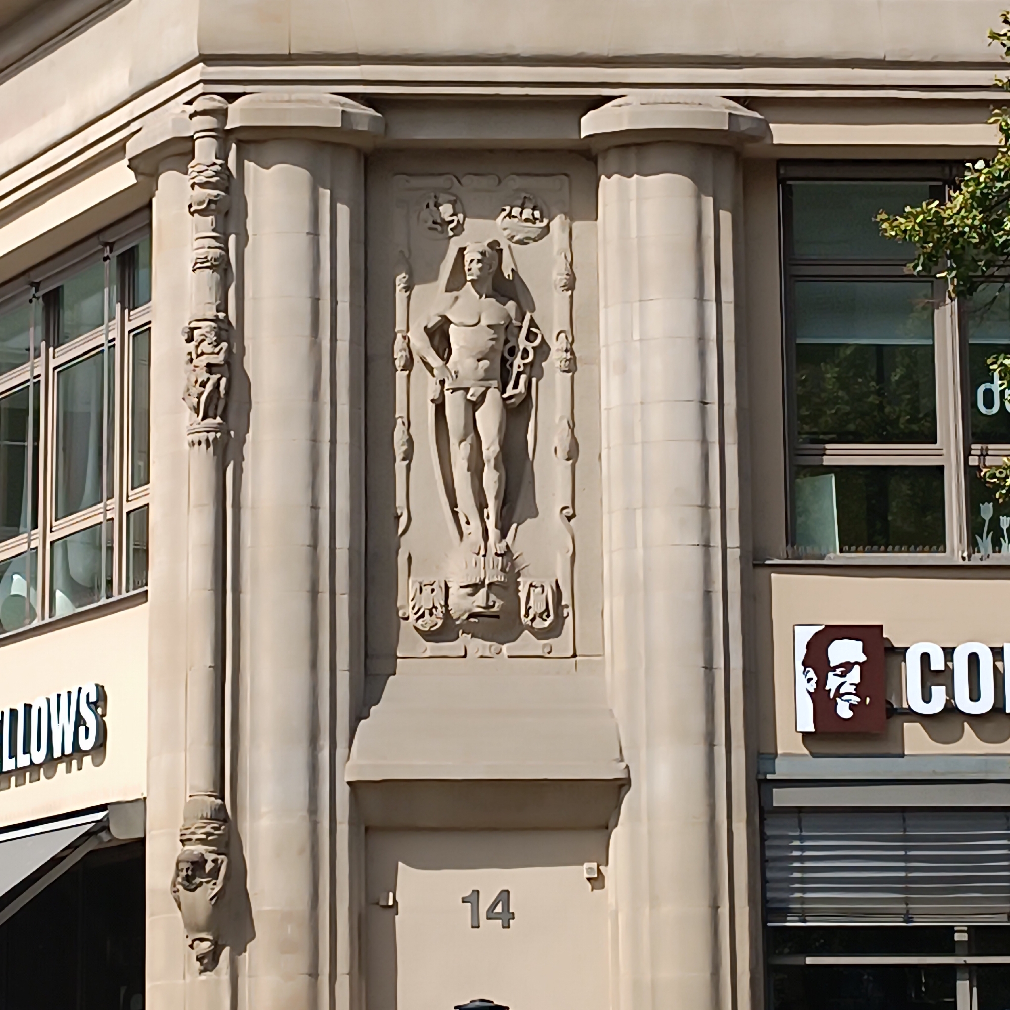 The image shows a corner of a building in Frankfurt, Germany, featuring architectural ornamentation and signage. Prominently displayed on the building is a bas-relief sculpture of a male figure in classical attire, holding a staff and a caduceus. To the right of the sculpture, a sign with "CO" is visible, accompanied by a stylized portrait of a man's face. Below this, a window with blinds is seen. To the left of the sculpture, a window with a reflective surface and a sign reading "LLOWS" is partially visible. The building's facade is made of light-colored stone, with columns and decorative elements. A large numeral "14" is affixed to a section of the wall below the sculpture. The overall impression is of a historical or ornate building in an urban setting.