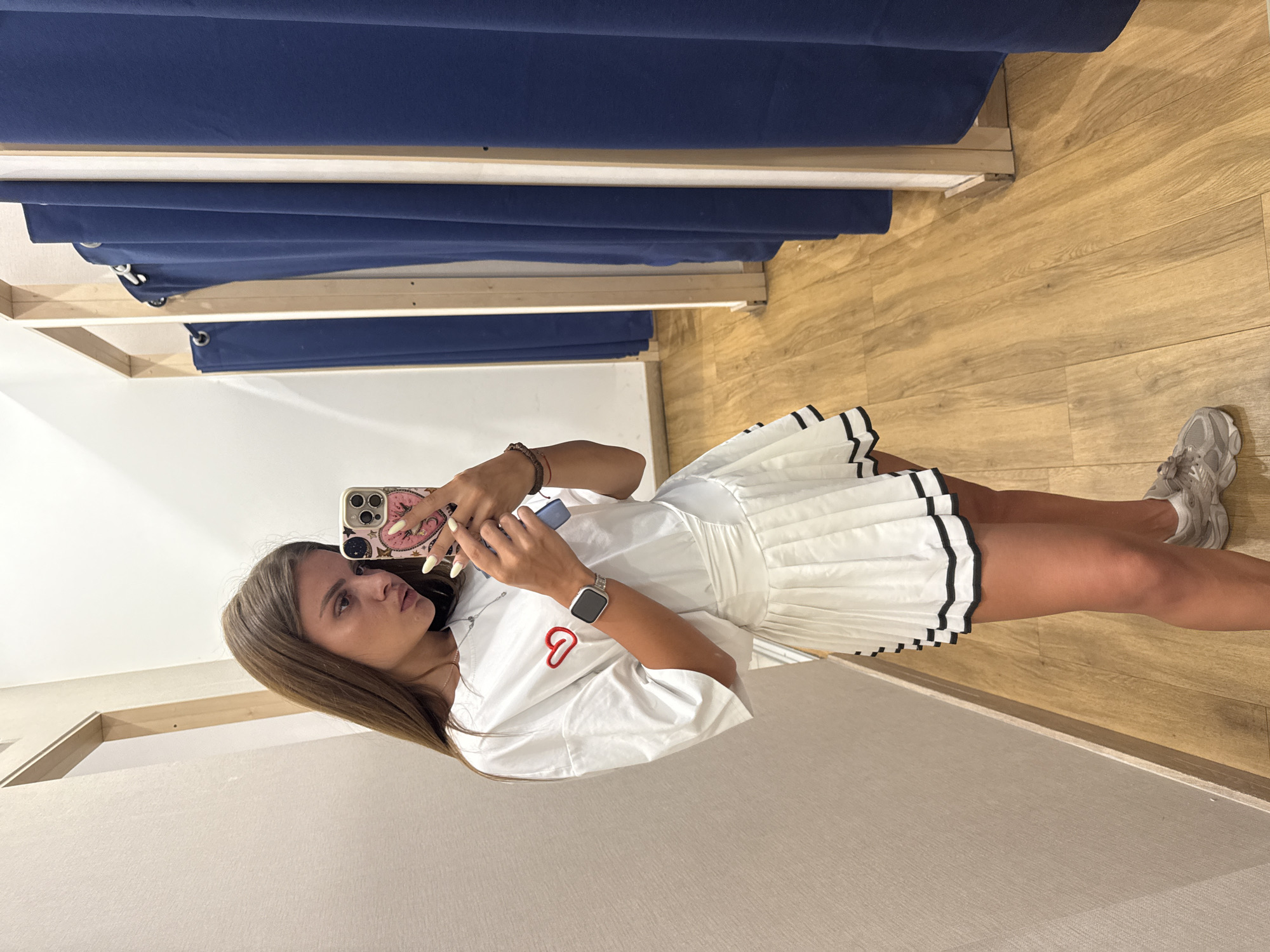 In a fitting room with light wood floors and beige walls, a young woman stands and takes a mirror selfie. She is wearing a white t-shirt with a red heart graphic on the left chest, a white pleated skirt with black trim, a silver smartwatch on her left wrist, and grey sneakers. Her long brown hair is down, and she is holding a pink phone in her right hand. The fitting room has wooden frames and navy blue curtains that appear to divide separate stalls. The overall lighting is bright and even, suggesting an indoor environment.