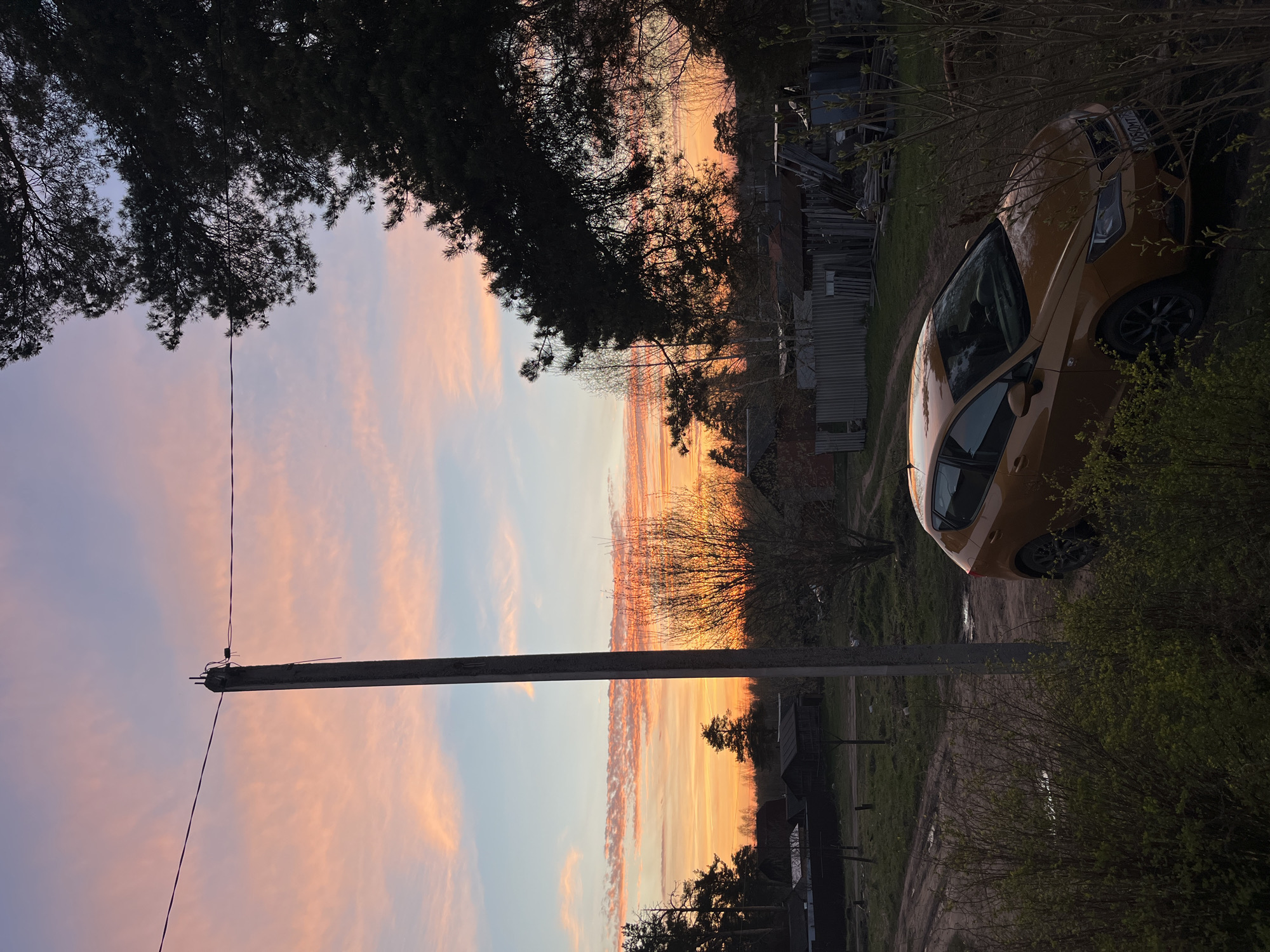 The image depicts a view in Razdolie, Russia, at dusk or dawn. The sky is a mix of orange, pink, and light blue. A utility pole stands vertically in the middle ground. In the upper right corner, an orange or yellow car is parked on a dirt path alongside some vegetation. In the background, partially obscured by trees, there are structures and sheds. The foreground and top of the image are framed by silhouetted tree branches.