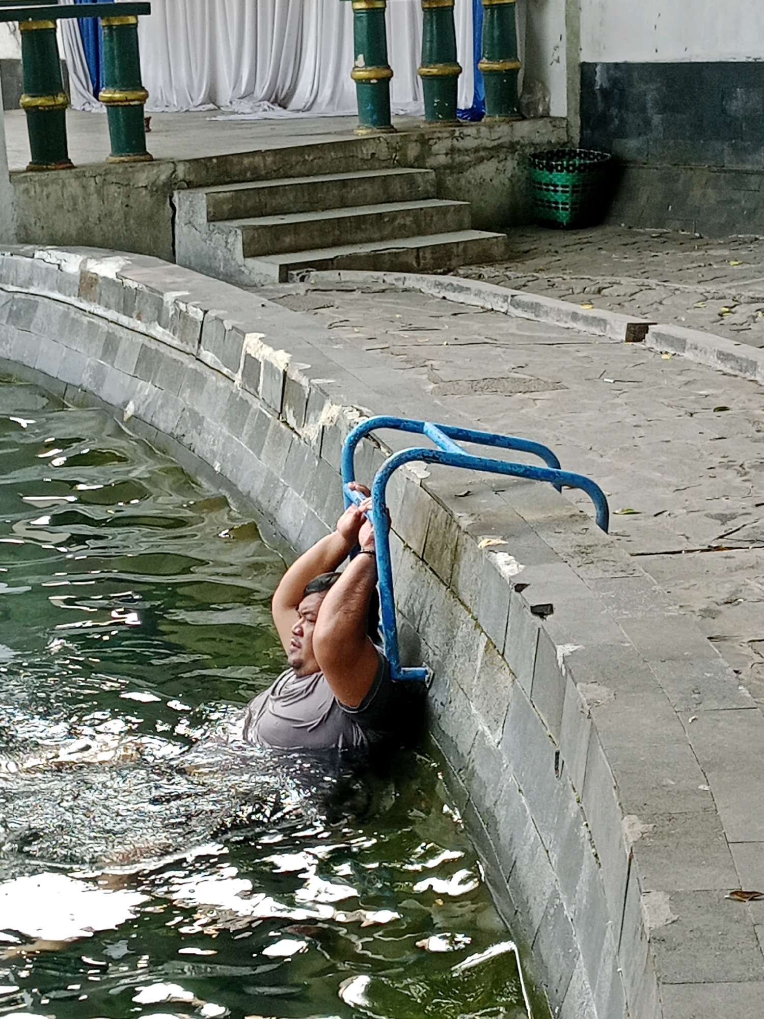 A man is holding onto a blue railing in a body of water with a stone edge. The man is partially submerged in the water, wearing a gray t-shirt. Behind him, stone steps lead up to a paved area with a green woven basket. Green and gold pillars with white drapes are visible in the background. This scene is located in Jentekan, Indonesia.