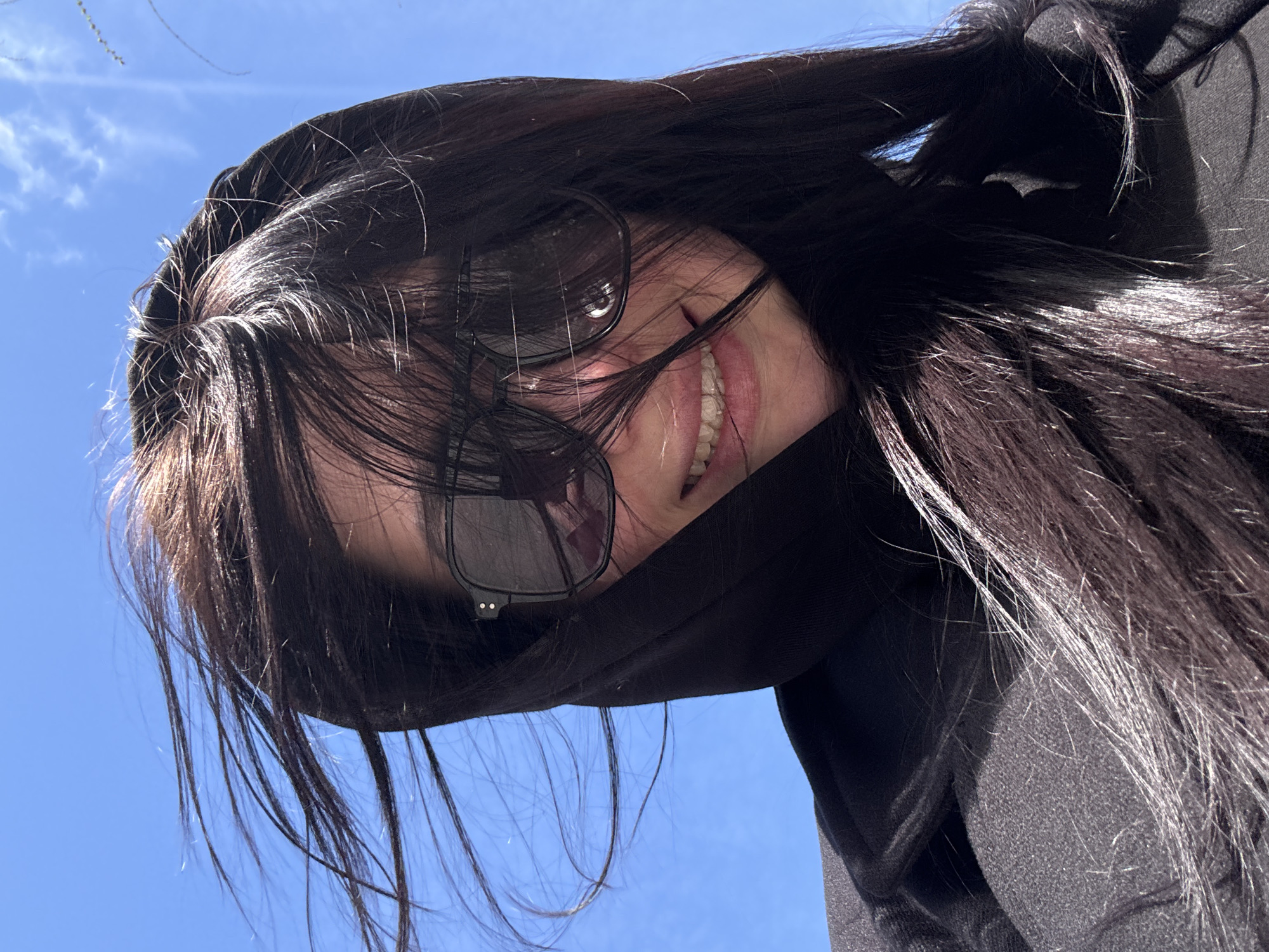 A woman with long, dark hair wearing sunglasses and a black hooded sweatshirt is looking down at the camera. The background is a bright blue sky. The location is unknown.