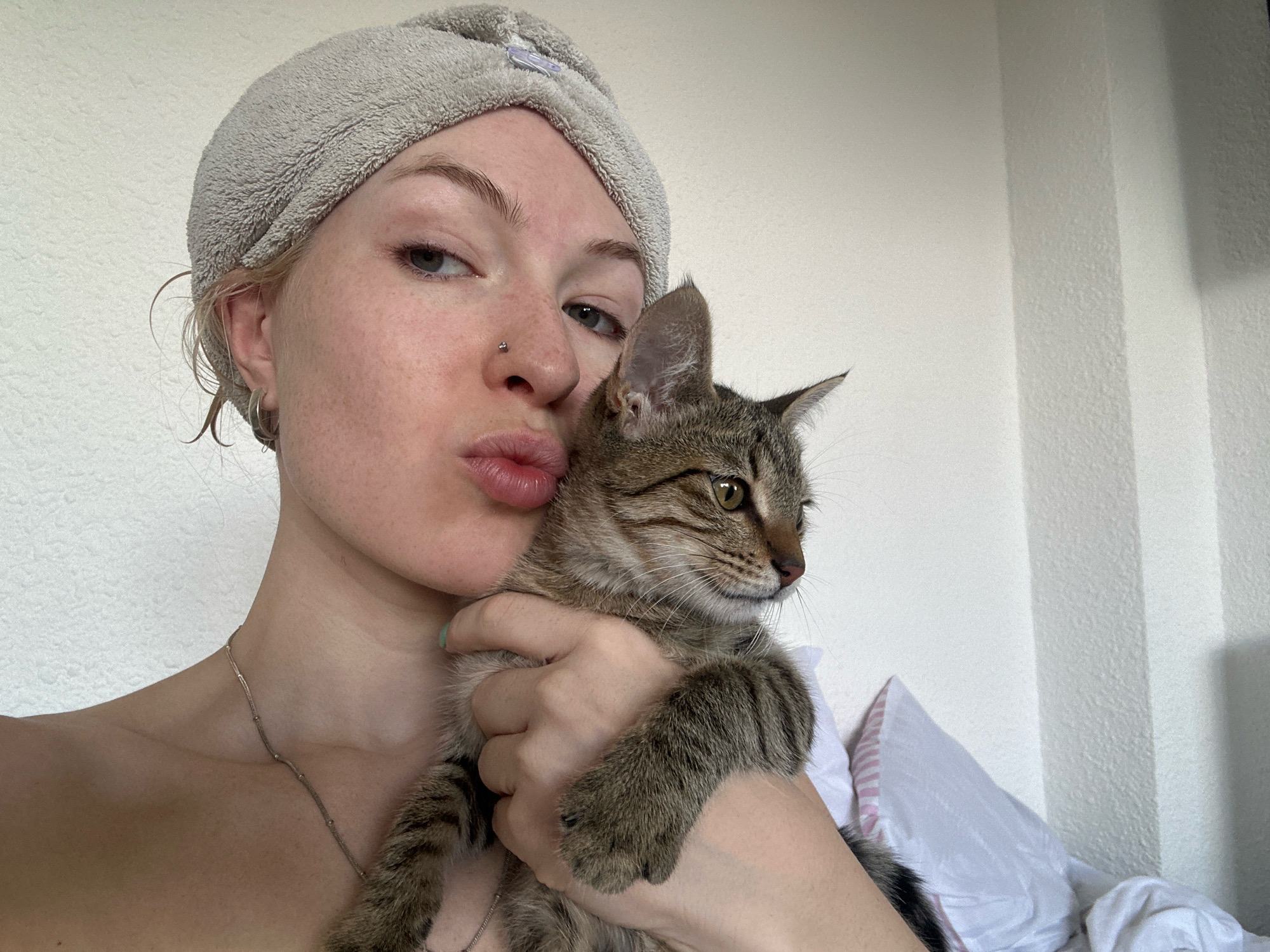 In a close-up, self-taken photograph, a young Caucasian woman with blonde hair wrapped in a grey towel and a nose piercing leans towards the camera with pursed lips. She is holding a small tabby kitten, which is looking to the right of the frame. The woman is wearing a silver necklace. The background is a white wall, possibly in an indoor setting. The lighting suggests it is daytime. There is no information in the image to suggest the location is Tbilisi, Georgia.