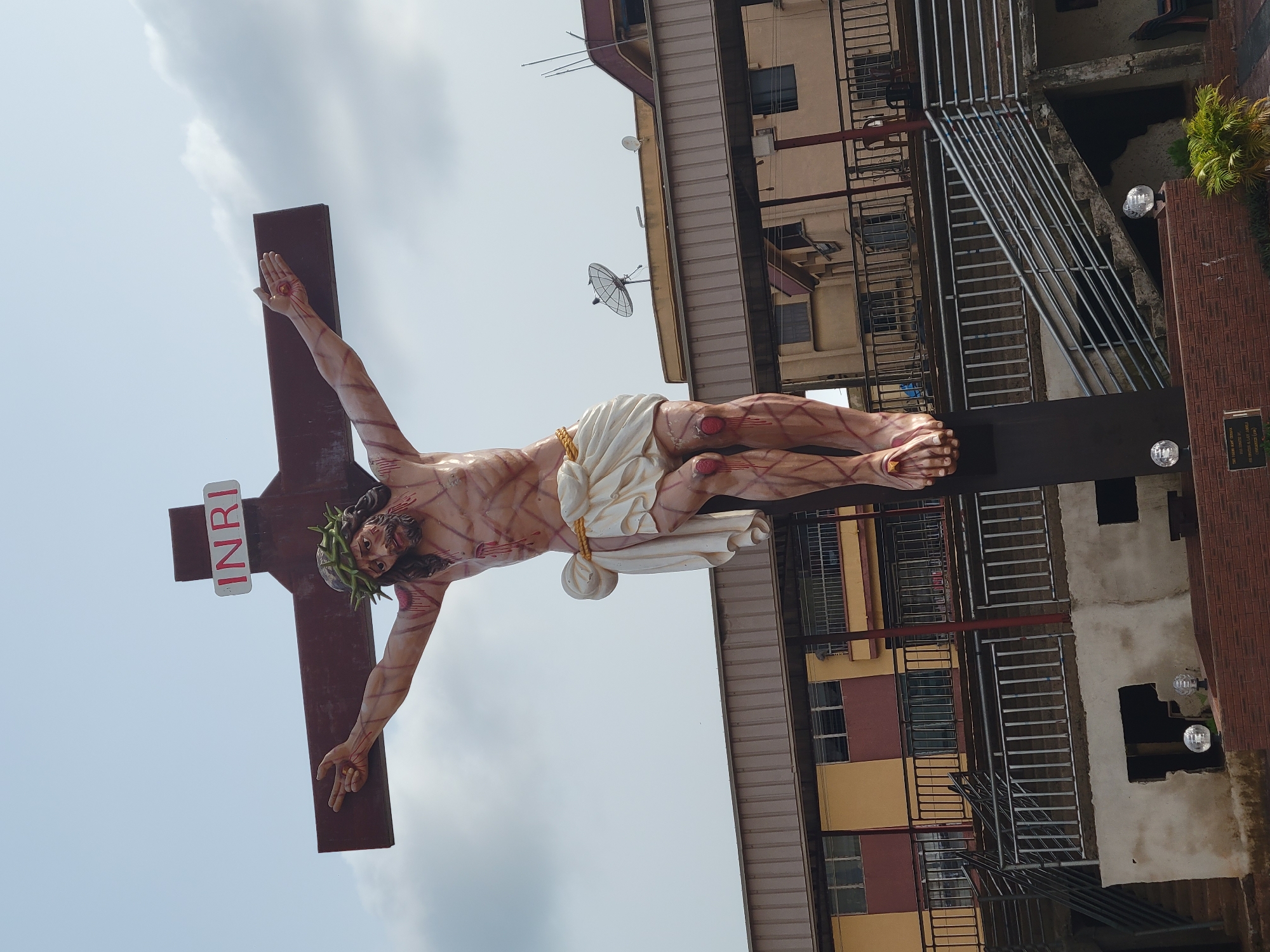 The image depicts a statue of Jesus Christ on the cross, bearing visible wounds, set against a background of buildings in Onitsha, Nigeria. The statue is positioned outdoors, under a cloudy sky. Buildings of multiple stories, constructed with concrete and painted in yellow and brown hues, occupy the background. Fire escape ladders and a satellite dish are attached to one of the buildings. A portion of a brown brick wall and foliage are visible on the right side of the image.
