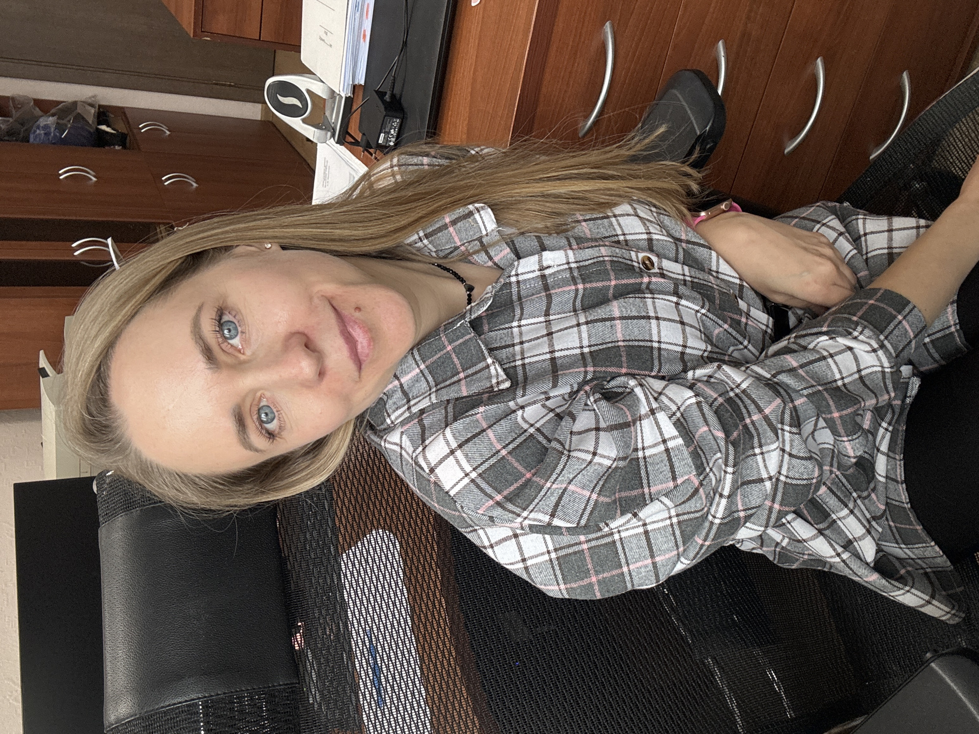 A woman with blonde hair and blue eyes is sitting in a black mesh office chair. She is wearing a gray, white, and pink plaid flannel shirt. The chair is positioned in front of a wooden desk and cabinet set. On the desk, there is a computer monitor, a stapler, and some papers. A pair of gray shoes is visible near the cabinet. The location is likely an office in Chaikovsky, Russia.