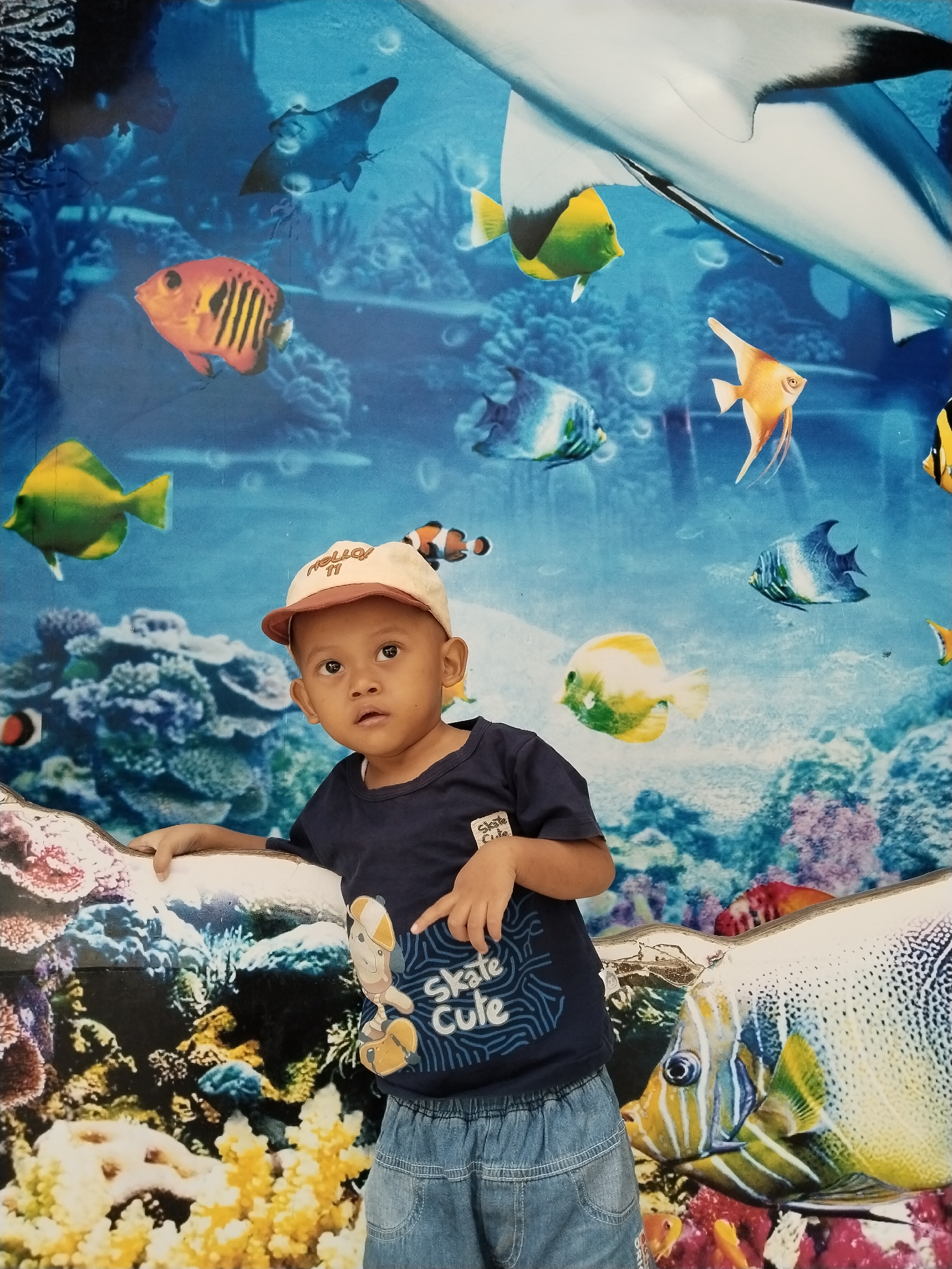 A young boy wearing a blue t-shirt and a cap stands in front of a mural of an underwater scene with various fish and coral. The boy is looking slightly upwards and to the left. The mural depicts a shark, angelfish, clownfish, and other marine life. The background of the mural is a bright blue, representing water. The foreground of the mural shows colorful coral formations. The scene appears to be indoors, possibly in a public place given the nature of the mural, and could be located in a recreational facility or a shop in Kecamatan Pati, Indonesia.