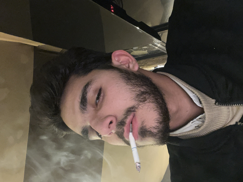 The image features a young man with dark, thick hair and a full beard, captured from the chest up, smoking a cigarette indoors. His head is tilted slightly to his right, and his dark eyes gaze upwards and to the side. He is wearing a dark, possibly black, jacket or hoodie, with a light-colored, ribbed collar visible beneath it, suggesting an underlying shirt.
A lit cigarette is held between his lips, with visible smoke curling upwards and to the left of his face, indicating he is actively smoking. The cigarette itself is white with a distinct blue band near the filter. The background of the image is dark and largely indistinct, suggesting an indoor setting with low or artificial lighting, possibly indicating evening or nighttime. The man's expression is neutral, appearing calm or contemplative. The scene captures a moment of quiet activity. The photograph was taken in Barza Municipality, Syria.