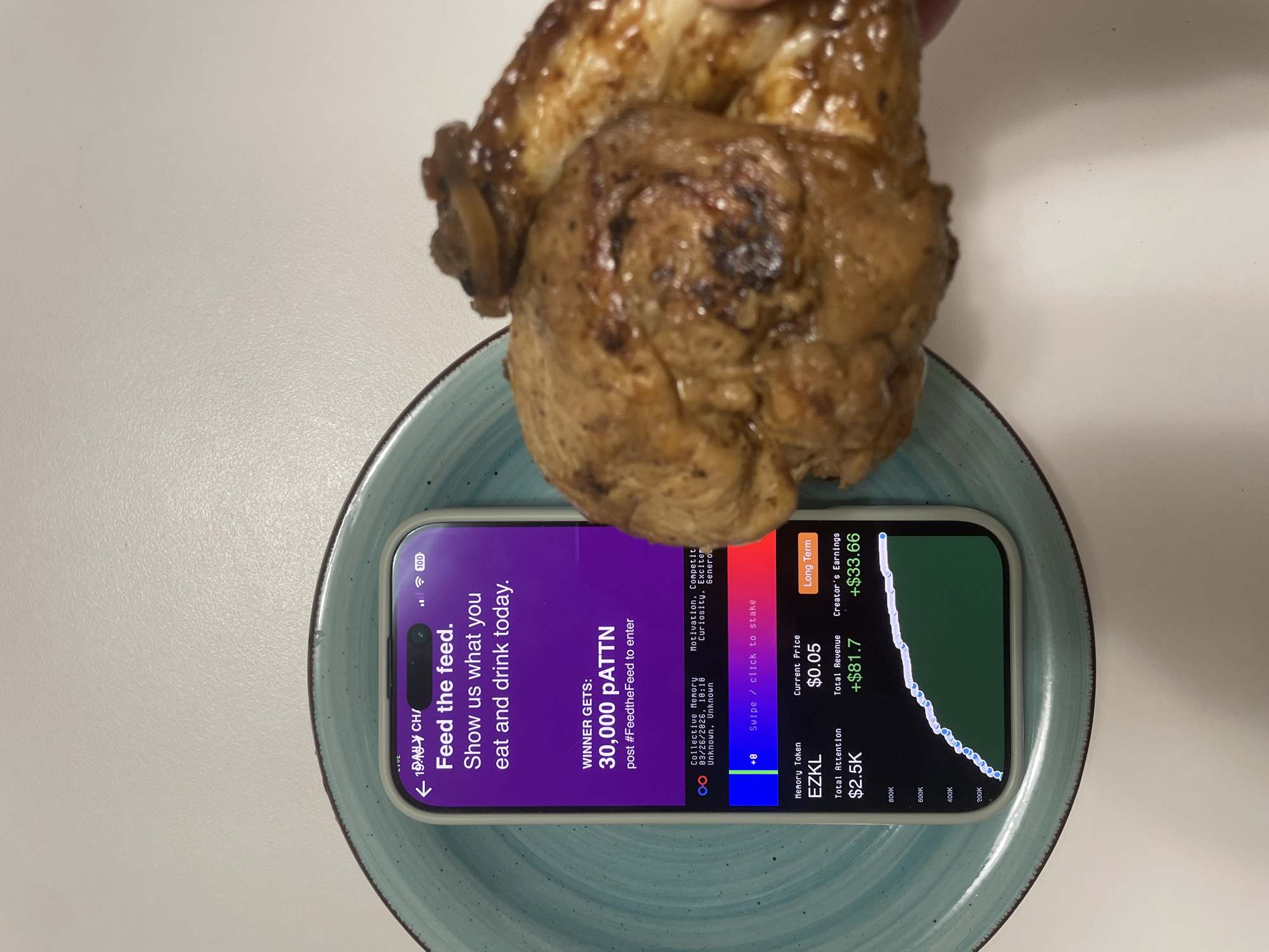 The scene, located in Budva, Montenegro, presents a close-up, top-down view of objects arranged on a light-colored, textured surface. A light-colored smartphone, likely an iPhone, is positioned on a round, teal-colored ceramic plate. The smartphone's screen displays an application with a dark interface, featuring a prompt: "Feed the feed. Show us what you eat and drink today," and details about a contest offering "30,000 pATTN." Financial metrics are visible, including "Memory Token EZKL," "Current Price $0.05," "Total Attention $2.5K," "Total Revenue +$81.7," and "Creator's Earnings +$33.66," along with an upward-trending green line graph. The phone indicates 100% battery charge. To the right of the smartphone, a human hand holds a piece of glazed, brown cooked meat, consistent with a portion of chicken or pork, which exhibits a bone and a mushroom cap garnish. The meat is positioned partially over the edge of the teal plate.