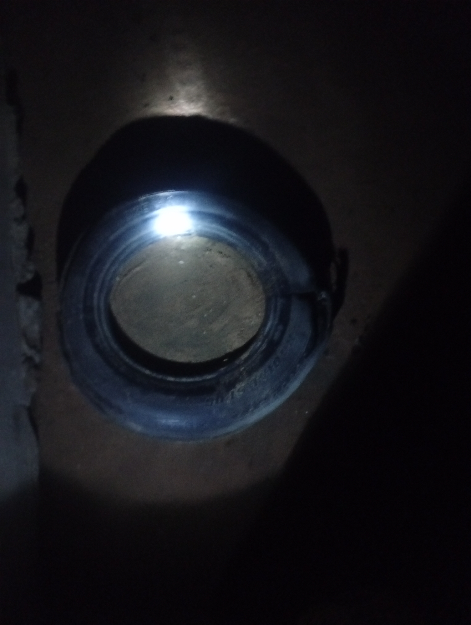 A close-up, high-angle shot depicts a black tire lying flat on a rough, dirt-like surface. A bright light source, possibly a flashlight, is aimed at the center of the tire, illuminating the inside opening and casting a strong glare. The tire's tread is visible along the outer edge, and some subtle markings or text appear on the sidewall. To the left of the tire, a rough, textured, greyish wall or surface is partially visible, creating a shadowed contrast. The majority of the background is dark, with the light from the source creating a dramatic spotlight effect on the tire. No people or specific actions are discernible. The environment appears to be outdoors or in a dimly lit, unfinished space.