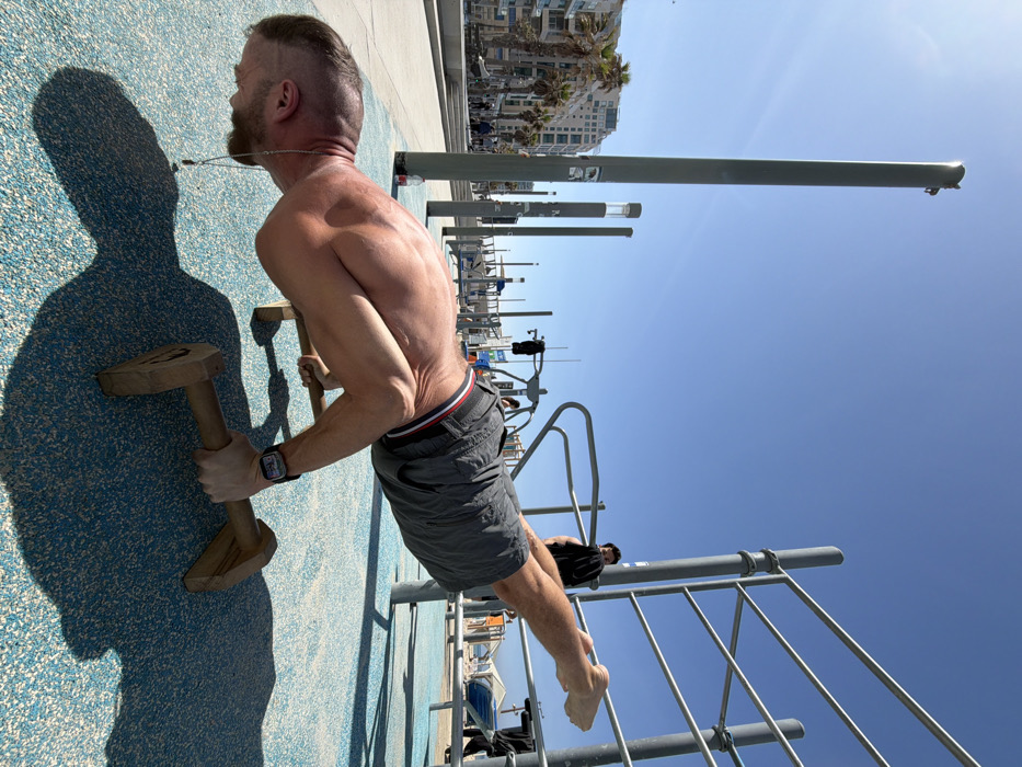 The image depicts a shirtless, muscular man performing a calisthenics exercise, likely a planche or a deep pseudo-planche push-up, on two wooden parallettes. He is positioned horizontally, body parallel to the ground, with his head facing downward and bare feet extended behind him. He wears grey shorts with a red, white, and blue stripe on the waistband and a dark smartwatch on his left wrist. A thin necklace with a dark pendant is visible.
The scene takes place outdoors on a sunny day with a clear, bright blue sky, casting distinct shadows from the man and equipment. The ground beneath him is a speckled blue, textured surface, consistent with a rubberized outdoor gym floor. In the immediate background, to the right, are sections of grey metal calisthenics equipment, with another man in dark clothing partially visible behind them. Further in the background, a sandy beach with sunshades or tents is visible, leading to what appears to be the Mediterranean Sea. To the left, a paved promenade with pedestrians is visible, flanked by numerous tall palm trees and multi-story buildings, typical of beachfront architecture. Several tall, grey utility poles with lights or cameras are positioned in the mid-ground.
This location is identified as an outdoor gym facility along the Tel-Aviv promenade (Tayelet) in Tel-Aviv, Israel, specifically near the city's beaches.