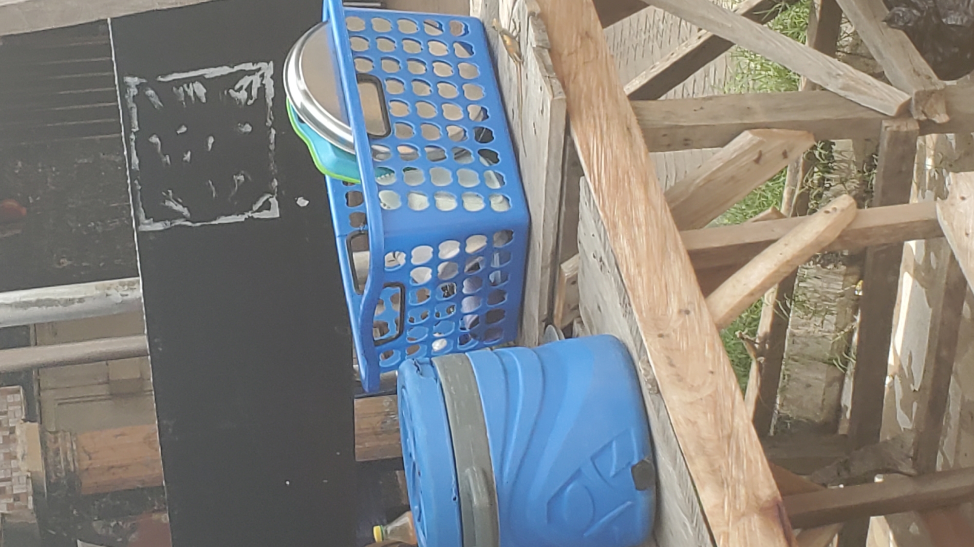 In Jalingo, Nigeria, a blue plastic water dispenser and a blue plastic basket filled with dishes are placed on a wooden table outdoors. The water dispenser is cylindrical with a textured design and a gray lid. The basket is rectangular with a lattice pattern and contains plates and bowls. A small lizard is visible on the table to the right of the basket. In the background, a dark wall with a white square marking and some textured elements is present. The environment appears to be a casual outdoor setting, possibly near a residential area or a market.