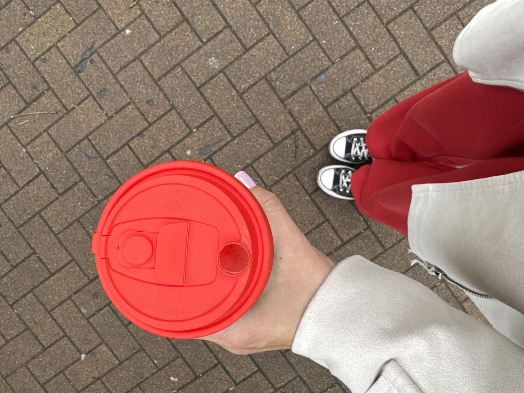 The image shows a top-down view of a person holding a red coffee cup with a plastic lid. The person is wearing red sweatpants, black and white Converse sneakers, and a light-colored jacket. They are standing on a brick walkway. The cup lid has a warning about hot contents written on it. The location in Sochi, Russia is not discernible from the image provided.