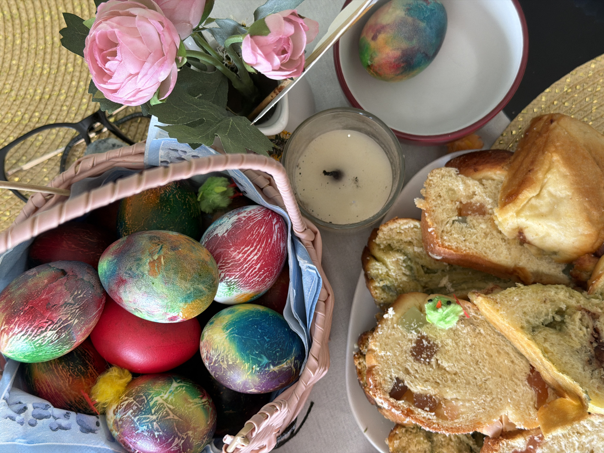 The image depicts an Easter celebration scene. A basket filled with dyed Easter eggs, some with marbled patterns, is visible on the left, along with a small yellow chick. Pink rose flowers in a pot sit adjacent to the basket. Nearby is a lit candle in a glass container. To the right, there is a plate of sliced bread with raisins, some pieces decorated with green icing. An Easter egg in a dish is present. Eyeglasses and a golden woven mat are also present in the background. There is no indication of the location, Bretton, United Kingdom in this picture.