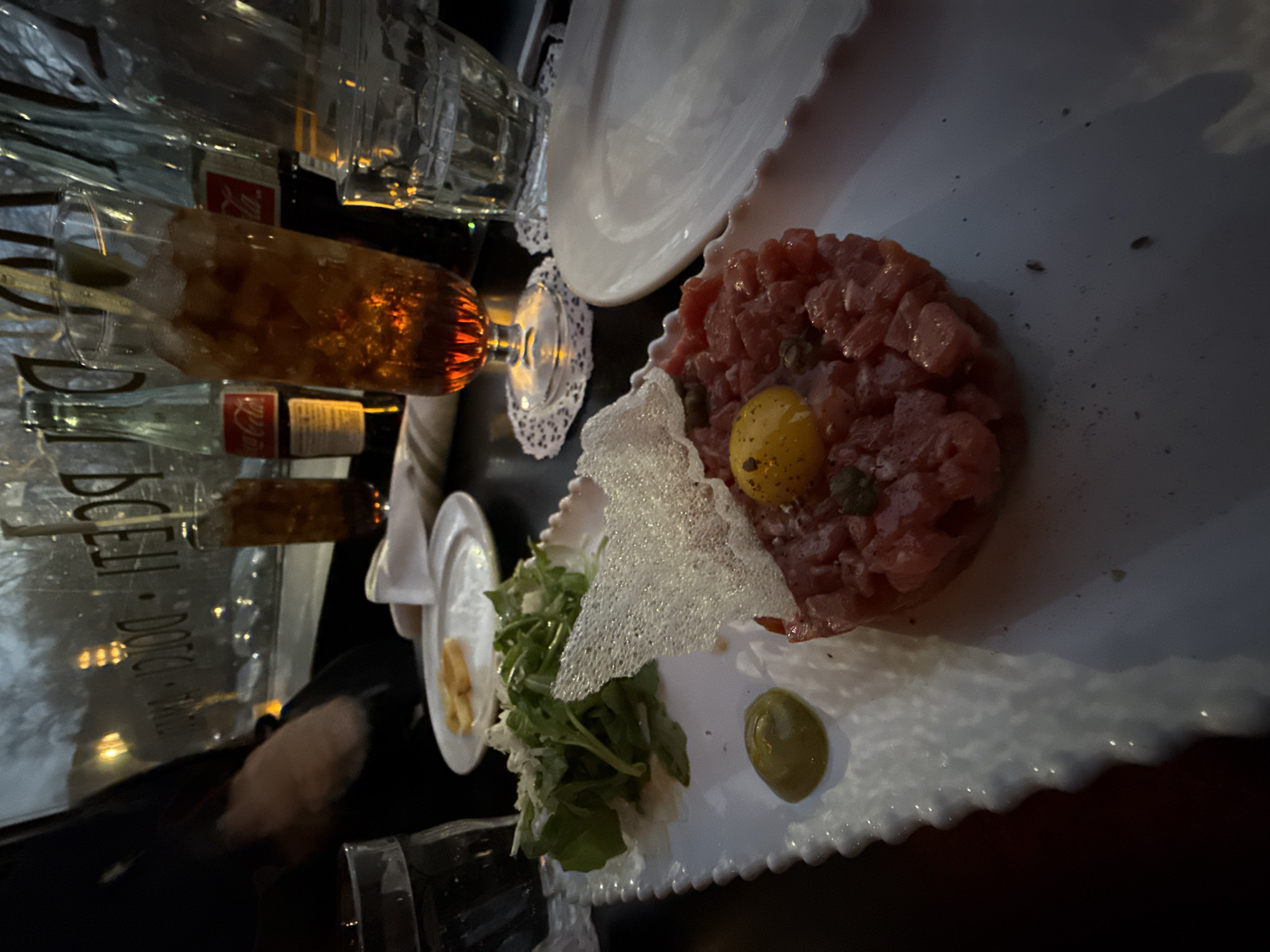 The image depicts a dining table within Docenti Pizzeria & Bar, located at 230 Park Avenue, New York, NY, United States of America. In the foreground, a white rectangular plate with a scalloped edge holds a dish of steak tartare, topped with a raw egg yolk, scattered capers, and a delicate, white, bubbly crisp. To the left of the tartare are a small pile of arugula salad and a dollop of olive-green sauce. A smaller white plate with two small, toasted bread pieces is visible behind. On the table, a tall glass containing an amber, ice-filled beverage with a straw stands on a white doily, positioned in front of several dark glass Coca-Cola bottles, some bearing red "Coca-Cola" labels. A clear glass of water is also present on the left. The background features a large window with reversed lettering, clearly displaying "DOCENTI" and "PIZZA," indicating the establishment's name. A blurry arm and hand are partially visible on the far left, suggesting a person is seated at the table. The lighting is dim, indicative of an evening or indoor setting.