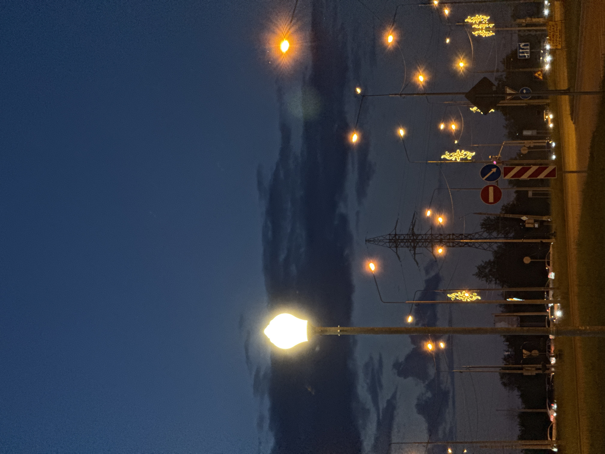 The image shows a night scene in a city. The sky is a deep blue, with dark clouds visible. Street lights illuminate the scene. Power lines and pylons are visible. A road runs alongside the scene, with street signs. Some lights are adorned with decorative patterns.