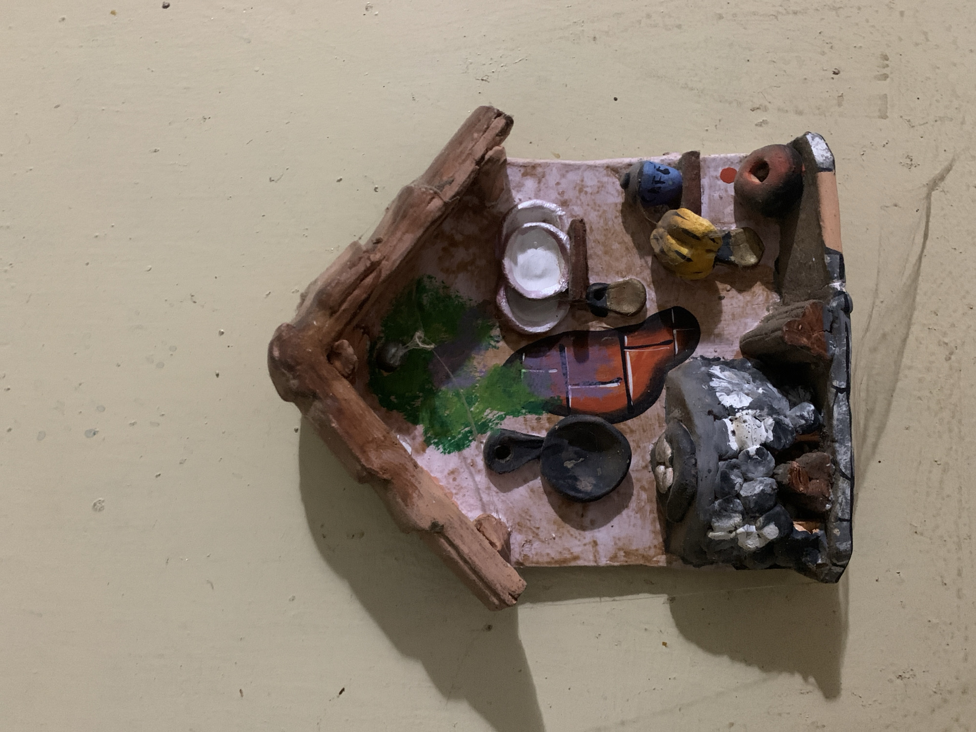 This media file captures a handcrafted miniature diorama mounted on a light beige wall. The diorama is shaped like an open-sided house or shed, constructed from rough, dark brown material resembling branches or carved wood.
Inside the miniature setting, the base is a light pinkish-purple hue. On the left side, there's a patch of painted green, suggesting grass or foliage. Towards the center, two small white bowls or plates are stacked, and nearby lies a black miniature frying pan with a handle. To the right, a rustic stove or hearth is depicted, made of grey and black materials with white accents, suggesting ash or embers, and containing reddish-brown elements within. Other small, colorful objects are scattered in the upper right portion, including a blue cylindrical item, a yellow, somewhat bird-like figure, and a reddish-brown ring-shaped object.
The scene portrays a simple, rustic cooking area or a small, humble dwelling. The image is taken from a slightly elevated angle, looking down into the diorama, which itself is oriented diagonally on the wall. No specific activity is taking place as it is a static art piece. There is no visible text. The location context provided is Santa Ana, Venezuela. The lighting suggests an indoor setting, with shadows cast by the diorama onto the wall.