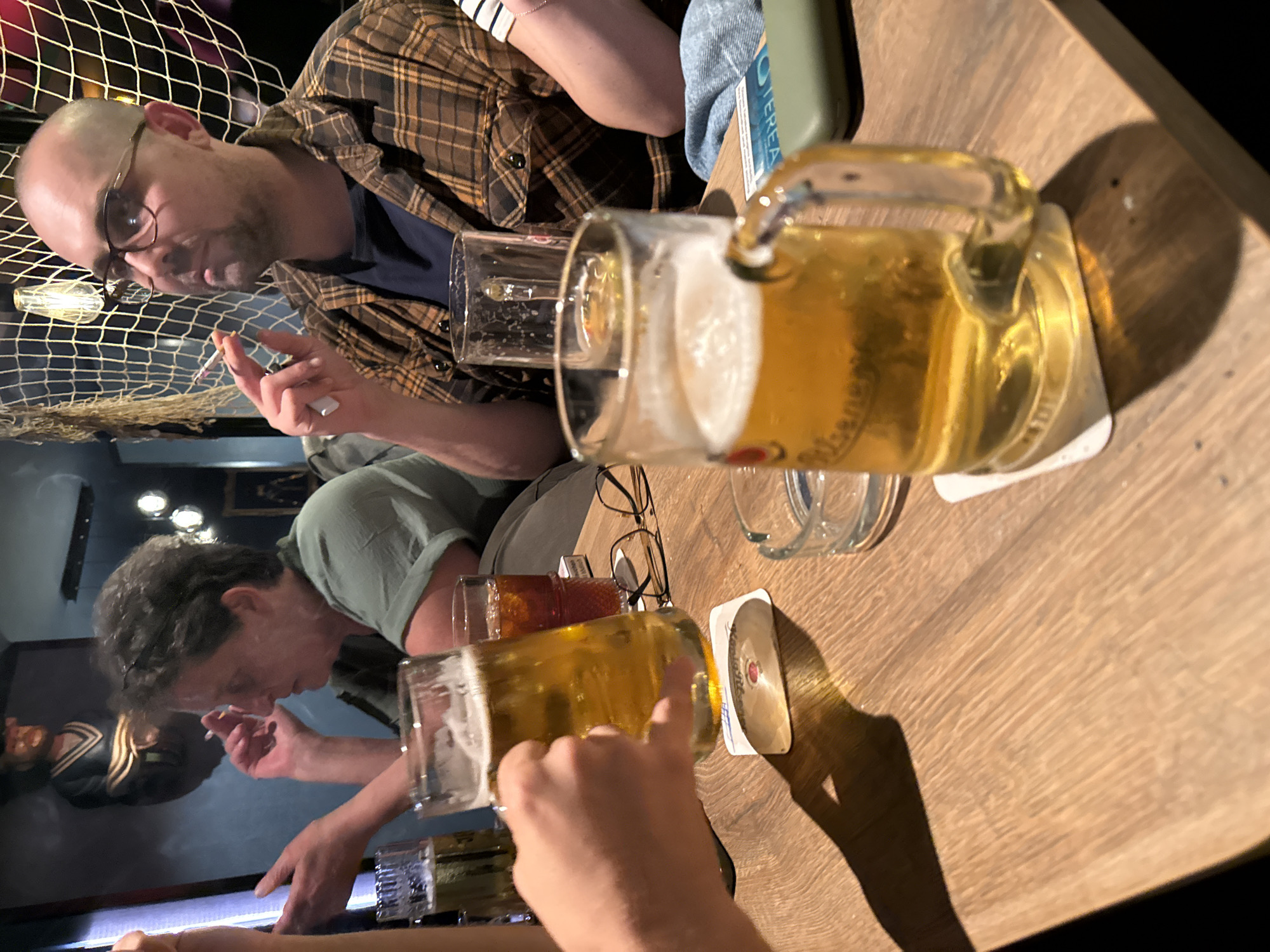 In a dimly lit establishment with nautical-themed decor, possibly a bar or restaurant in Langeoog, Germany, four individuals are seated around a wooden table laden with beverages. Two men and two women are present, though not all are fully visible. One man, with glasses and a bald head, wears a plaid shirt and holds a cigarette. Another man, with short grey hair, also holds a cigarette. Two glasses of beer are prominent on the table, one being held by an unseen hand, its condensation indicating it is cold. A glass with a darker liquid, possibly iced tea or soda, also sits on the table, alongside a cigarette lighter and ashtray. The background features a fishing net draped over a light fixture and a framed artwork depicting a person in nautical attire. The lighting is warm and atmospheric, casting shadows across the table.