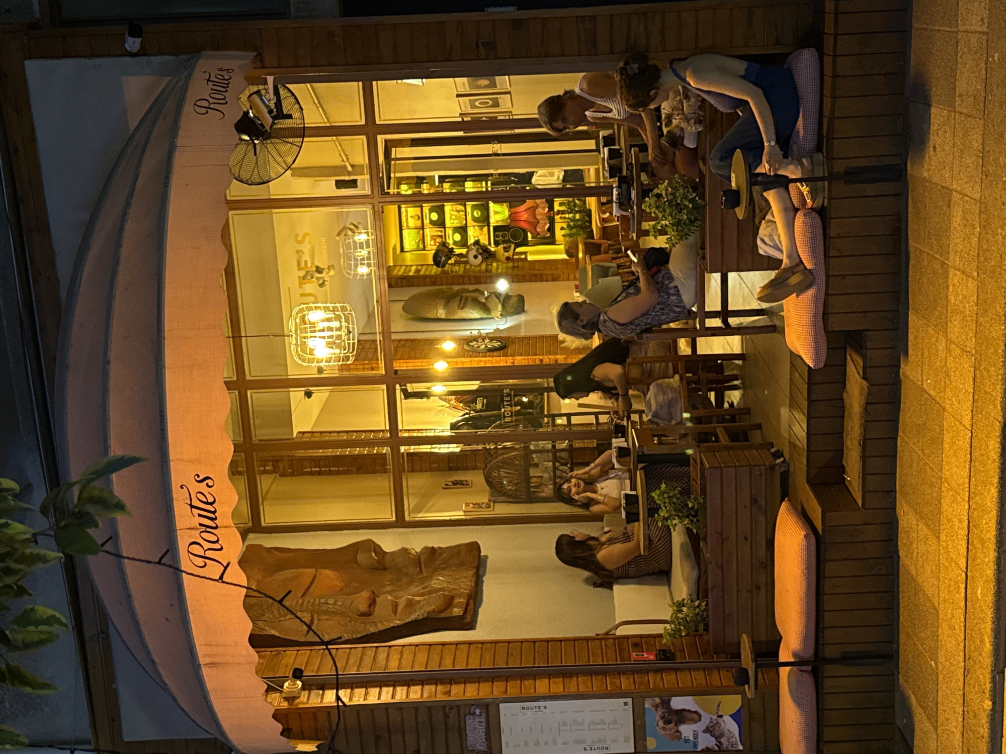 This image shows a street-level view of "Route's", a cafe located in Kadıköy, Turkey, at night. The cafe has a prominent orange awning with the cafe's name in cursive script. Large glass windows reveal the warm interior, where several people are seated at tables. In the foreground, on the outdoor seating area, three women are visible. One woman is seated on a cushioned bench, looking at her phone. Another woman, also on a cushioned bench, is facing away from the camera, with her legs extended. The third woman is seated at a small table, looking at her phone. Wooden planters with greenery are placed along the outdoor seating area. A large, carved wooden mask is mounted on the wall to the left of the cafe's entrance. The street in front of the cafe is paved with yellowish-orange interlocking blocks, illuminated by streetlights.