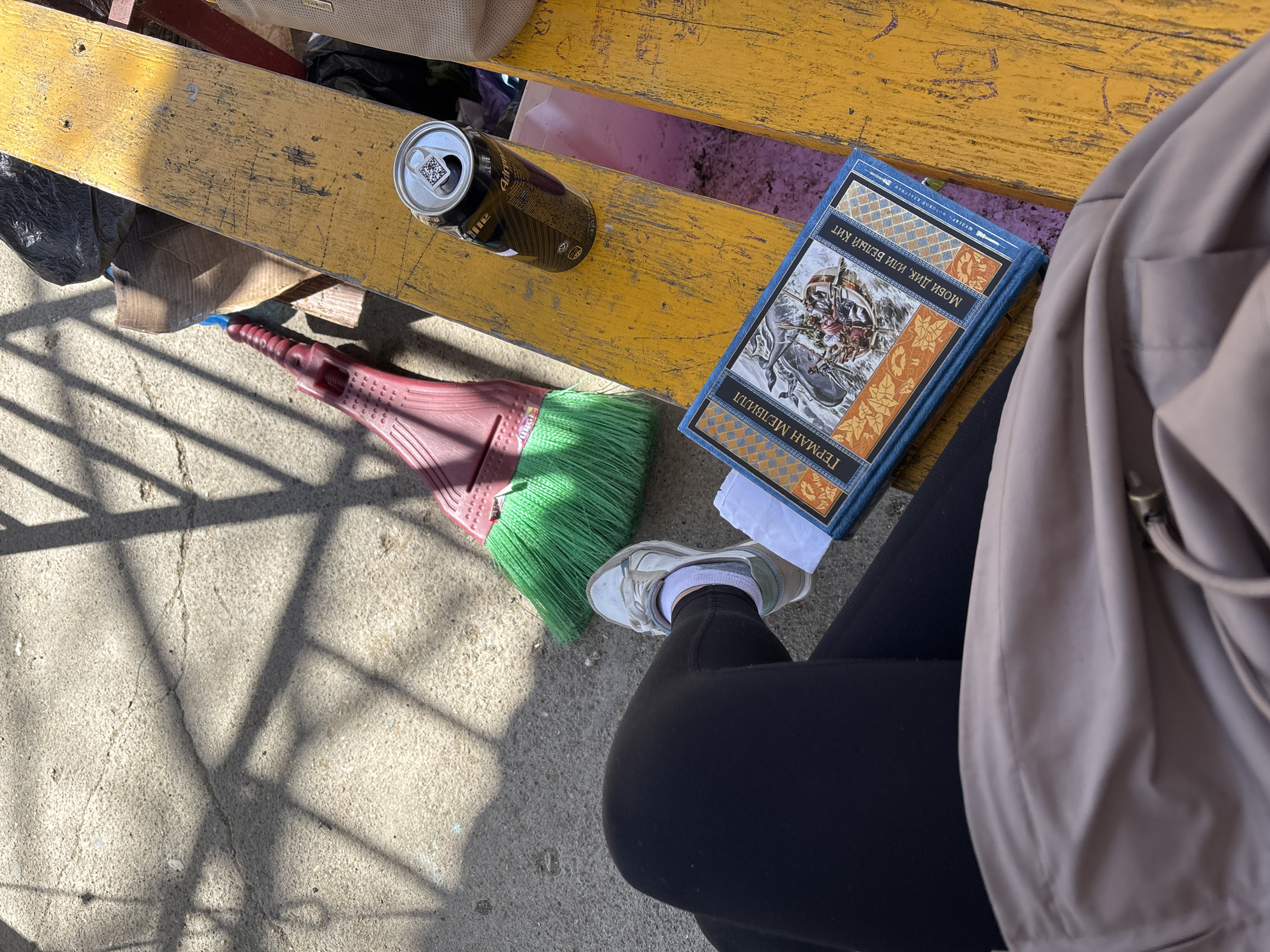 Captured from an elevated, high-angle perspective, the image presents a scene on what appears to be an old, yellow-painted wooden bench, likely situated outdoors. The setting is likely Sochi, Russia, based on the Cyrillic text on the book.
Several items are placed on the bench: a half-empty can of dark beer, a book with Cyrillic text on the cover, and a push broom with a red handle and green bristles. A white tissue is placed on the seat next to the book.
The book features a cover illustration with a whale. Part of a person is visible, wearing black leggings and a white running shoe. An adjacent beige jacket or bag suggests someone is seated on the bench. The background surface is concrete with a geometric shadow pattern.