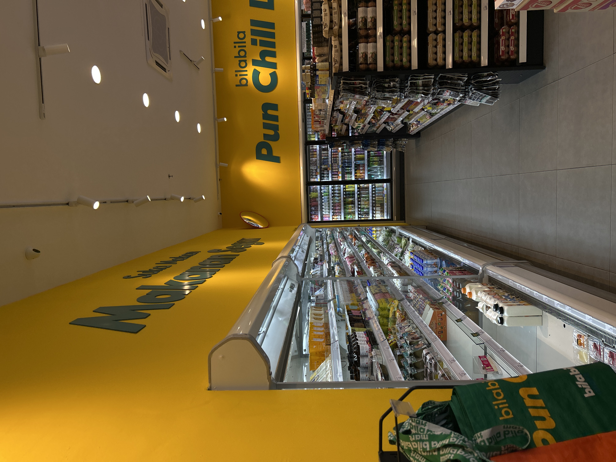 This appears to be an interior shot of a grocery store or supermarket, likely in Subang Jaya City Council, Malaysia, based on the writing on the walls and bags. The interior features refrigerated aisles stocked with various food and beverage items. The walls are painted a bright yellow with signage in black lettering. A grocery cart with green reusable bags is visible in the foreground. The store is brightly lit with ceiling lights. The floor is tiled and clean.