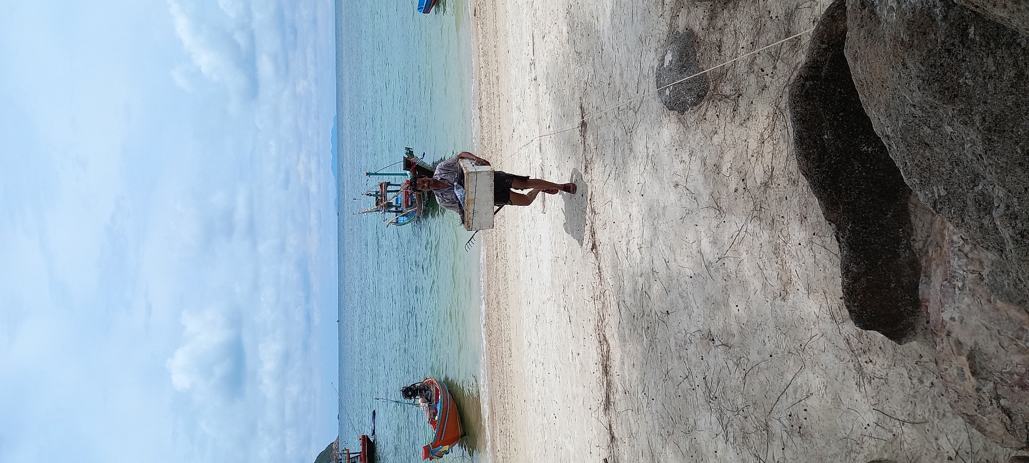 A man carries a white object on the beach in Baan Chaloklum, Thailand. The man, wearing a light-colored patterned shirt and dark shorts, is on the sandy beach with several fishing boats in the turquoise water. A large rock formation is visible to the right. The sky is partly cloudy.