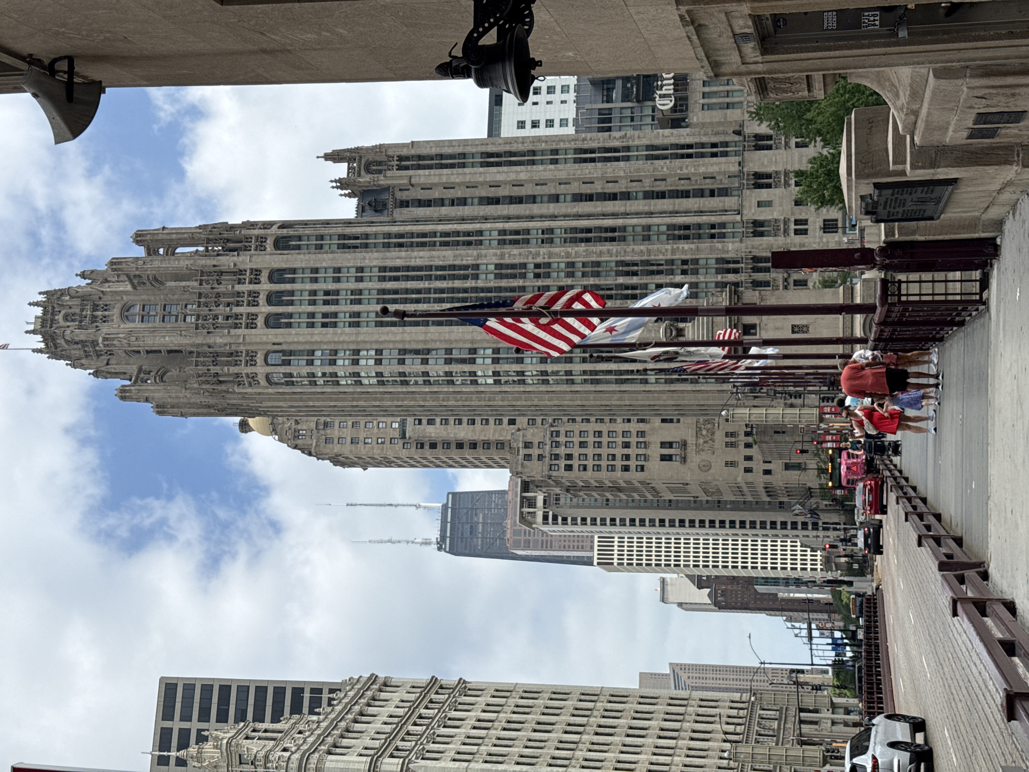 The image captures a view of Chicago's distinctive architecture, with several tall buildings dominating the skyline. In the foreground, an American flag and a Chicago city flag are visible, mounted on poles. The flags are partially unfurled, indicating a gentle breeze. A bridge or elevated walkway is present, with a railing lining the right side. Several people are walking along this walkway, some appearing to be tourists. One person in a red shirt and black shorts is particularly noticeable. Cars are seen in the distance, suggesting an urban environment with traffic. The sky is partly cloudy, hinting at a moderate weather day. The architecture of the buildings suggests a daytime setting, likely in the afternoon given the quality of light. The overall scene is one of a bustling city, with a focus on its impressive urban landscape and a sense of public space. No specific event is depicted, but the presence of people and vehicles suggests ongoing activity.