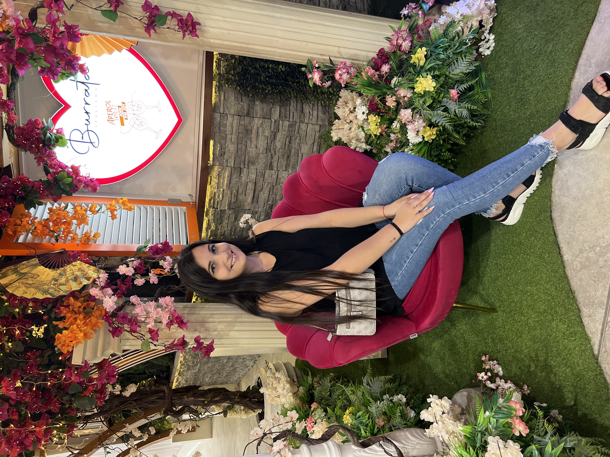 A young woman with long, dark hair smiles while sitting in a bright pink armchair. She wears a black sleeveless top, blue jeans with ripped hems, and black platform sandals. A small, light-colored crossbody bag hangs on her shoulder. The armchair is situated on a patch of artificial green turf, surrounded by lush floral decorations featuring pink, orange, and white flowers, as well as green foliage. Behind her, a stone wall serves as a backdrop. To her right, a large, illuminated heart-shaped sign displays the text "Burrata Italiana" and "Aperol Spritz Join The Joy." The setting appears to be an indoor or semi-outdoor space decorated for a festive or romantic ambiance. The location is identified as Sofia, Bulgaria.