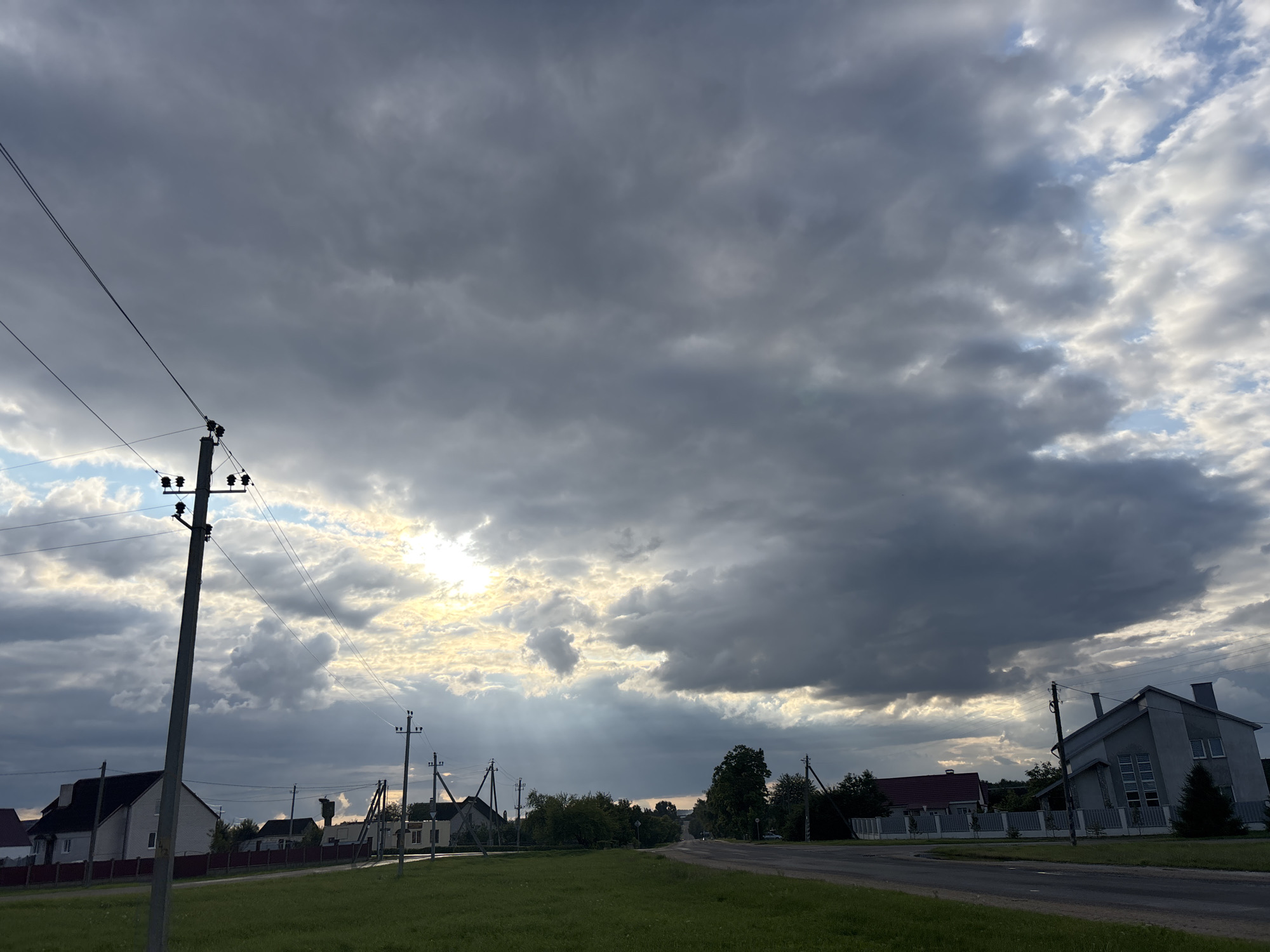 The image depicts a rural road in the city of Kapyĺ, Belarus, under a cloudy sky. A grey concrete utility pole with wires stands prominently in the foreground, slightly to the left of the center. To the left of the pole, a residential building with a dark roof is visible behind a red fence. Further down the road on the left, more houses with varying roof colors and designs are seen, interspersed with trees. On the right side of the road, a modern, multi-story building with a grey facade and a pointed roof is situated. Another house with a red roof is visible behind it. The road itself is asphalt, with a grassy shoulder and a wide expanse of green grass in the foreground. The sky is filled with dark, heavy clouds, but shafts of sunlight break through in the upper center of the frame, creating a dramatic effect. The overall scene suggests an overcast day, possibly before or after a rain shower. No people are present in the image.