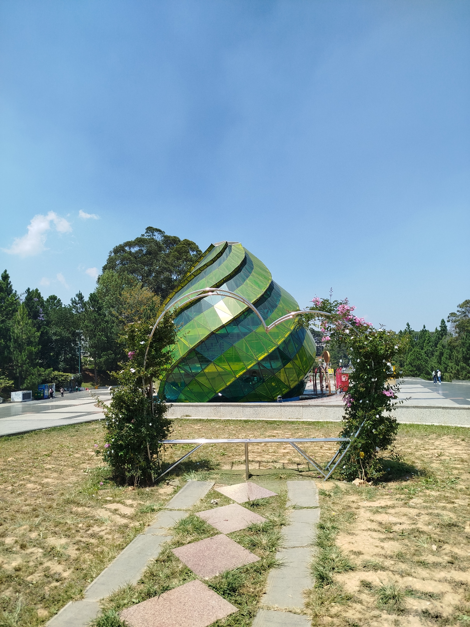 The image features the architectural structure of the Domaine de Marie Convent, situated in Da Lat, Vietnam. The green and yellow glass-covered structure has a conical shape. It is set against a backdrop of a clear blue sky and green trees. In the foreground, a path made of stone leads towards the structure, framed by two small trees. A low, silver metal frame surrounds these trees. The foreground is characterized by dry, brown grass. In the background, there are people walking on a paved area. The scene appears to be in a well-maintained park-like setting.