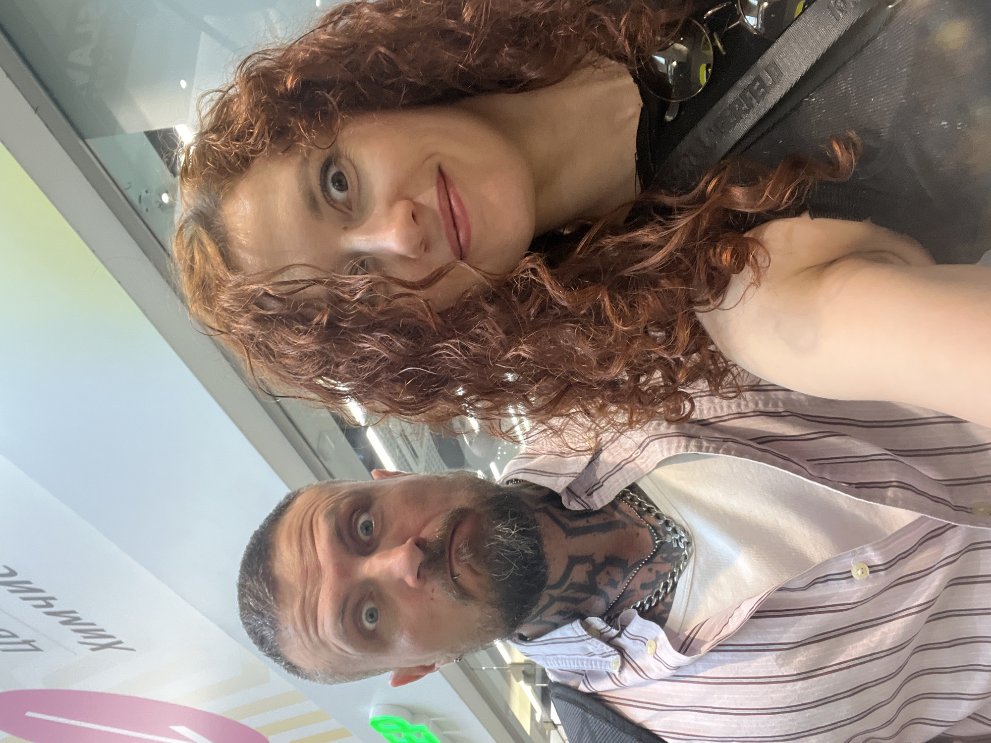 A selfie-style photograph, rotated counterclockwise, features a man and a woman posing together. The woman has curly red hair, light skin, and is positioned at the top of the frame with a slight smile. The man, located at the bottom, has a shaved head, beard, and visible neck tattoos. He wears a striped shirt over a white undershirt and a chain necklace. The background appears to be an indoor space with signage that includes Cyrillic characters and the words "ХИМИЧ" and "Ц." Based on the Cyrillic writing, it is likely the location is in Saint Petersburg, Russia.