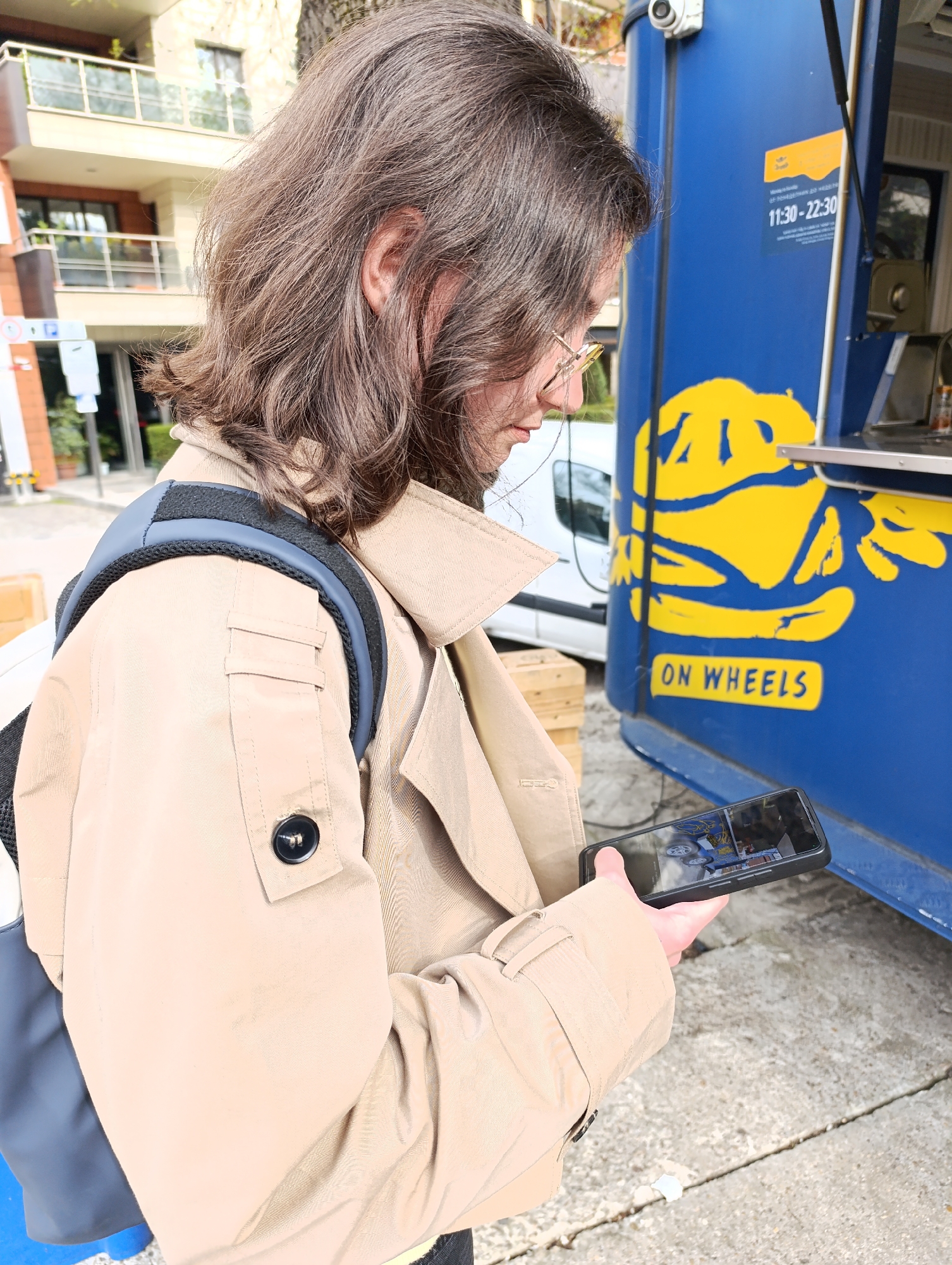 A person stands next to a blue food truck displaying a yellow logo and the words "ON WHEELS" with operating hours "11:30 - 22:30." The person is wearing a beige trench coat and a backpack, and is looking at a mobile phone. A building with balconies is in the background. The location is in Sofia, Bulgaria.