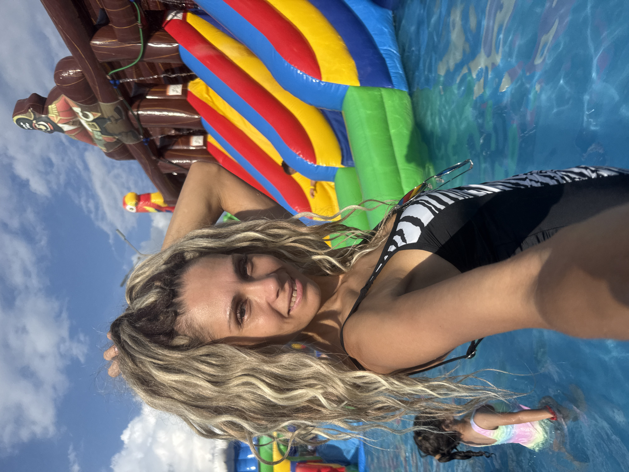 A woman with blonde, curly hair pulled into a ponytail takes a selfie at a water park with a large inflatable pirate ship and water slides in the background. She is wearing a black swimsuit with a white zebra print. The water is clear blue, and the sky is bright blue with white clouds. A young girl in a tie-dye swimsuit stands in the water in the background. The scene suggests a sunny day at an amusement park.