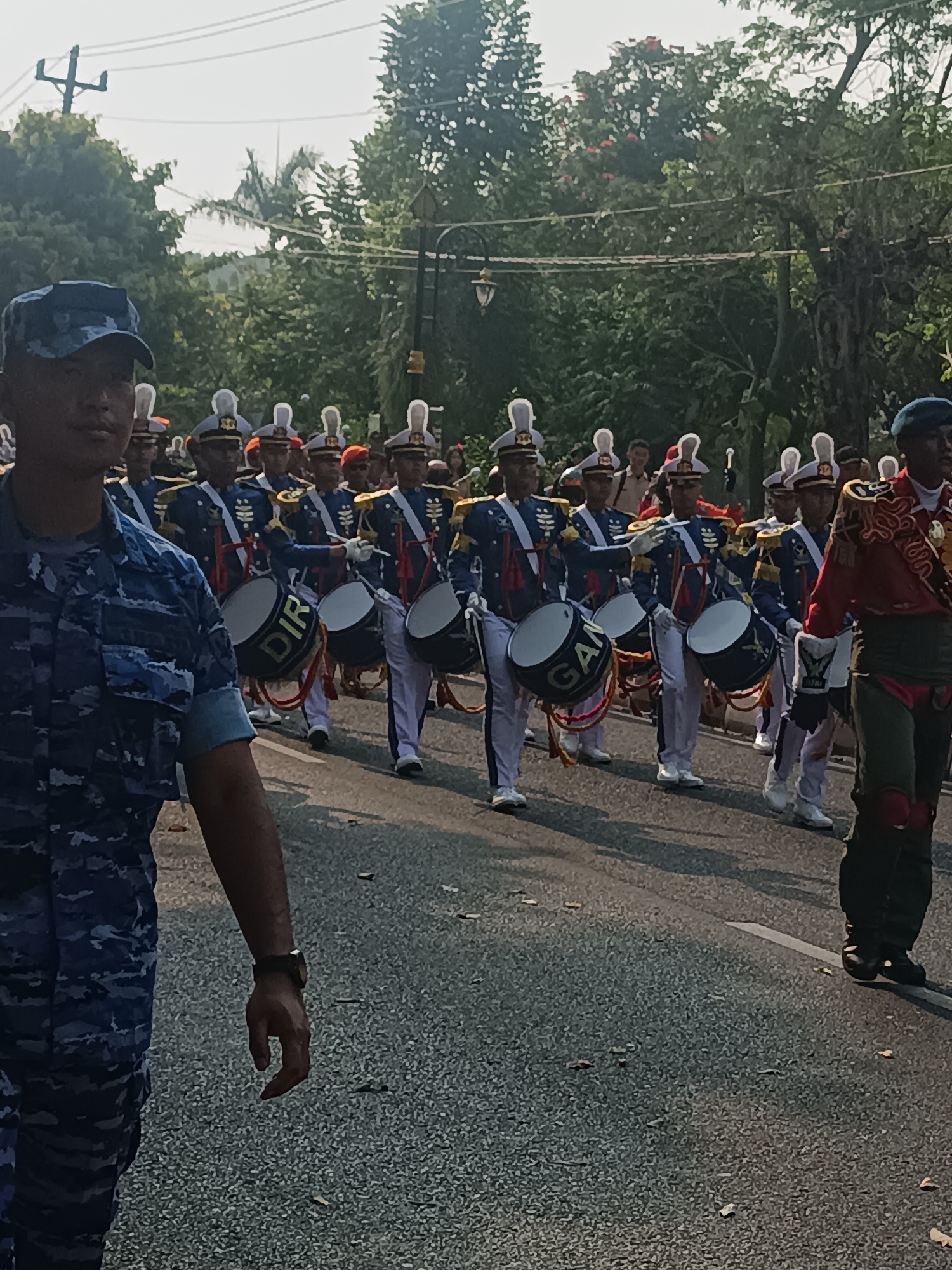 In Sleman, Indonesia, a marching band comprised of young men in ceremonial uniforms parades down a paved road. The band members, predominantly young men, are dressed in a variety of uniforms. Some wear dark blue uniforms with ornate gold trim and white pants, while others are in camouflage attire. Many are carrying or playing drums. In the foreground, a young man in a blue camouflage uniform walks forward, his left arm slightly bent and his right hand resting by his side. He wears a dark blue cap and a watch on his left wrist. Behind him, several rows of band members are visible. The band members in the middle ground are distinguished by their blue and gold uniforms, white pants, and tall white hats adorned with plumes. They are marching in formation, each holding a drum. The drums are predominantly black with white rims and some have writing on them, such as "DIR" and "GAM." The individuals in the background are less distinct, with some appearing to be spectators or part of the parade. The environment suggests a sunny day, with trees and foliage lining the street. Overhead, power lines are visible against a bright sky. The road surface is asphalt, with scattered leaves. The overall scene depicts a formal procession or parade in a city setting.