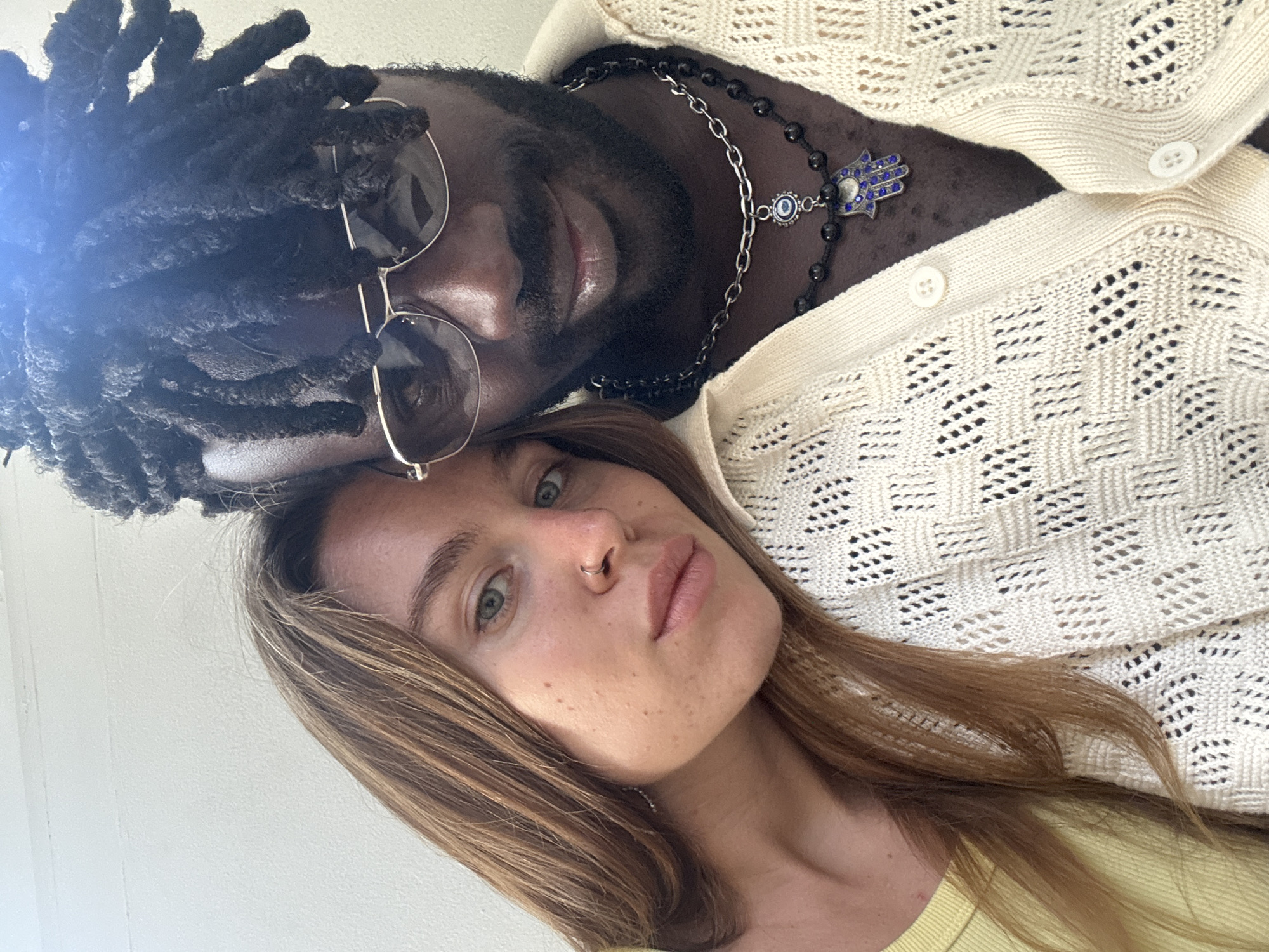 A close-up, selfie-style photograph shows two individuals, a woman and a man, positioned side-by-side. The woman, on the left, has light brown hair that falls over her shoulder. She is wearing a yellow top. The man, on the right, has dark dreadlocks and is wearing glasses and a cream-colored open cardigan over a dark shirt. He is wearing two necklaces, one with black beads and a pendant that appears to be a blue hamsa hand with an evil eye symbol, and another silver chain above it. The background is a plain, light-colored wall, suggesting an indoor setting. There is a bright light source, possibly a ceiling light, casting a glare in the upper right portion of the image. The image is focused on the faces and upper bodies of the two individuals, capturing a close and personal moment. There is no discernible location within the city of Mengwi, Indonesia.