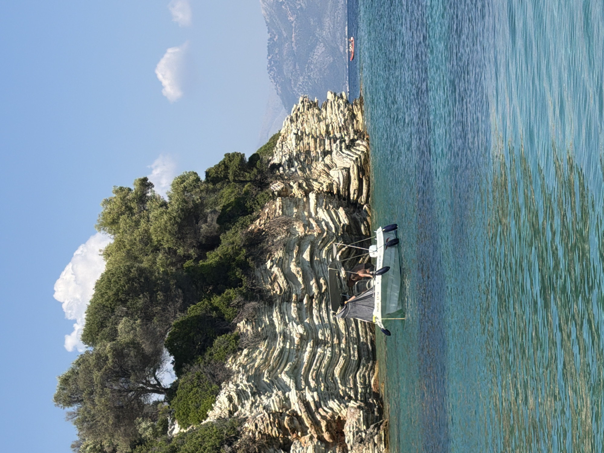In the Municipal Unit of Kastos, Greece, a small white motorboat with a canvas canopy is anchored in clear turquoise water, close to a layered, rocky cliff face covered in dense green foliage. A person, partially visible, is seated in the boat. The boat has several black fenders on its sides. In the distance, a larger red boat is visible on the sea, with hazy, mountainous terrain in the background. The sky is blue with a few scattered white clouds.