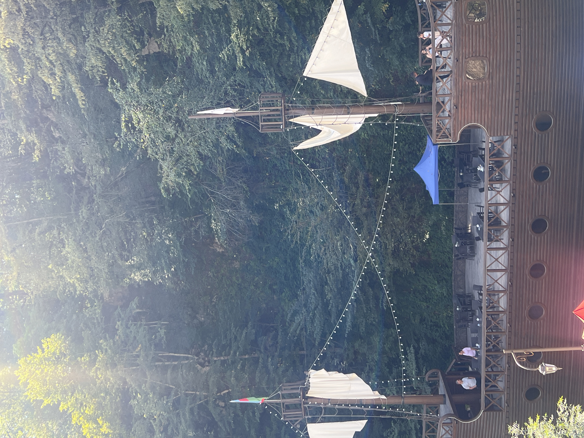 The image depicts a unique restaurant in Sarısa, Azerbaijan, designed to resemble a wooden pirate ship. The vessel is situated against a backdrop of dense, green forestation. The ship features two masts with white sails, one of which is topped with a flag. A walkway encircles the upper deck, where tables and chairs are set up for outdoor dining. Several patrons are visible on the deck, some seated at tables, others standing and conversing. A blue canopy tent is also present on the deck. The lower part of the ship shows a wooden hull with circular windows. In the foreground, a street lamp is visible. The lighting suggests it is daytime, with sunlight filtering through the trees.