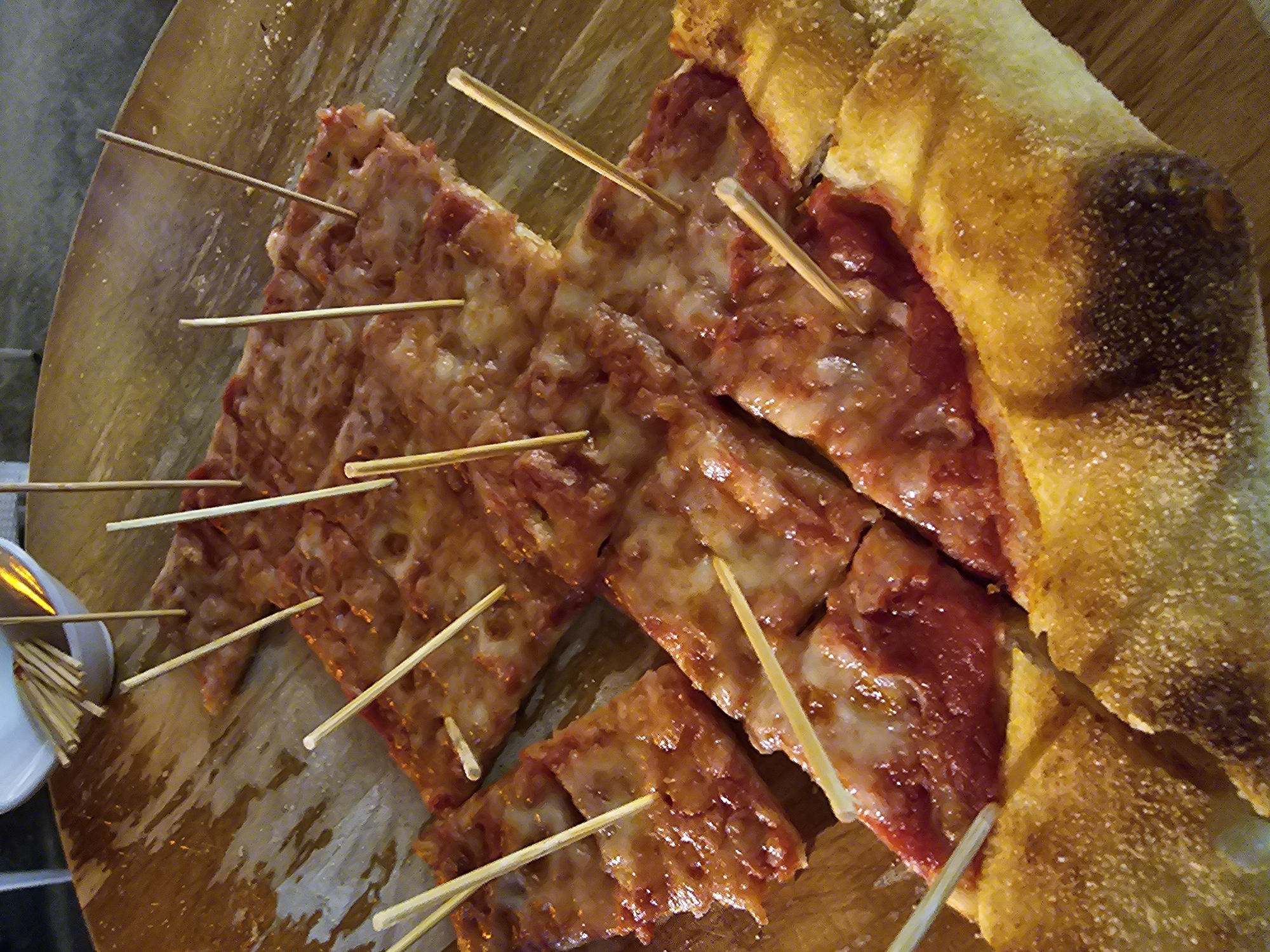 A high-angle shot depicts a cut, square pizza served on a wooden platter with numerous toothpicks piercing each slice. The pizza has a red topping, likely tomato sauce, and a lighter topping, possibly mozzarella cheese. The crust appears thick and golden. To the left of the platter is a small, white container filled with more toothpicks. The background is blurred. While the photograph does not provide conclusive evidence to pinpoint the exact location within the city of Denpasar, Indonesia, it is possible to infer that the pizza is being served in a restaurant or eatery located in Denpasar.