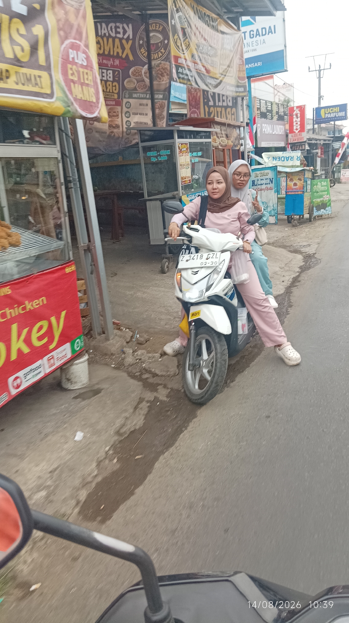 The image shows two women on a white scooter on a street in Tasikmalaya, Indonesia. The woman in the front is wearing a pink hijab and a pink outfit, and the woman behind her is wearing a light blue hijab and a light blue outfit. They are both smiling. The scooter has a license plate that reads "Z 3418 CZL". In the background, there are several street vendors with their stalls. One stall has a red banner with the words "Chicken Pokey" and logos for GoFood and Food. Another stall has signs that read "KEDAUNG MAX", "PLUS EST EH MANIS", and "GRAJI GADAI". The overall environment appears to be a busy street in a city. The timestamp on the image indicates it was taken on August 14, 2026, at 10:39 AM.