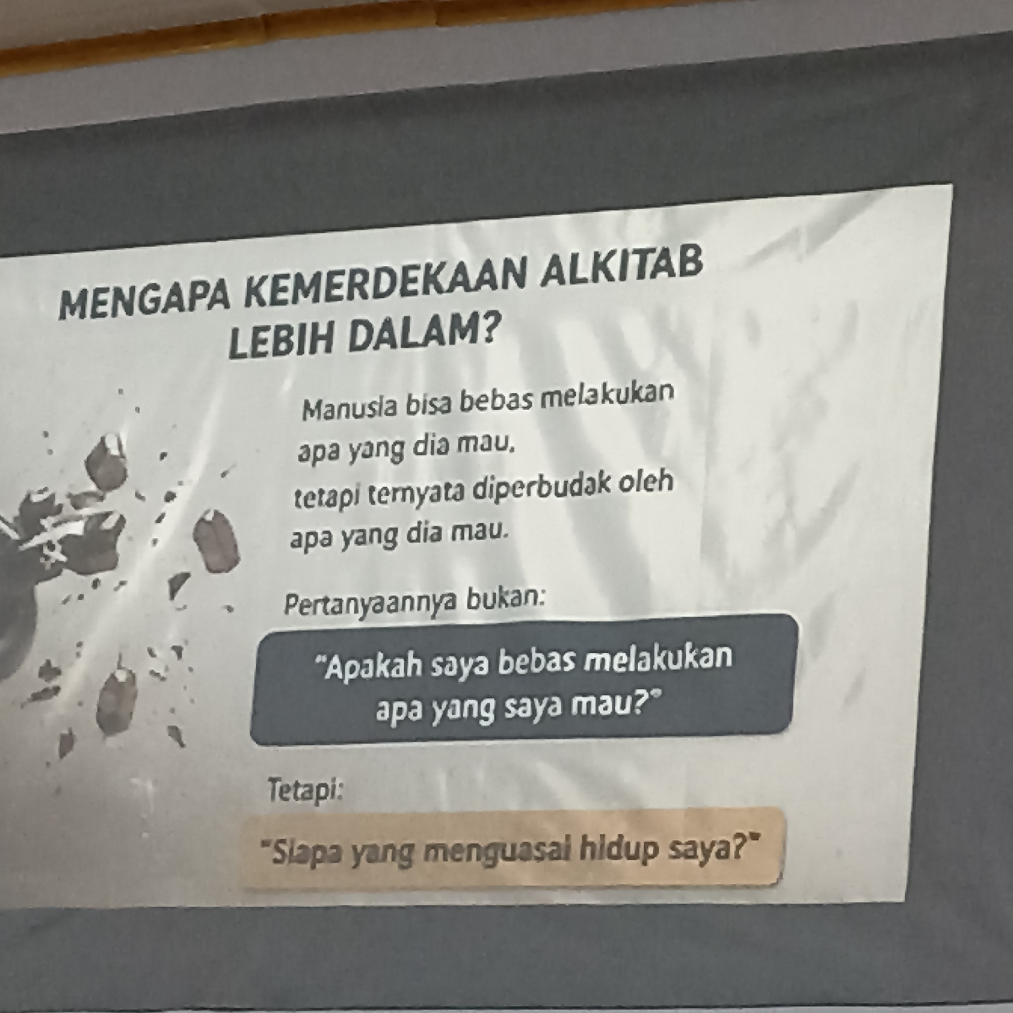 The image shows a poster with text in Indonesian. The title of the poster is "MENGAPA KEMERDEKAAN ALKITAB LEBIH DALAM?". Below the title, there is text that translates to "Humans can be free to do what they want, but it turns out they are enslaved by what they want." Following this, there is a question: "Pertanyaannya bukan: 'Apakah saya bebas melakukan apa yang saya mau?'" which translates to "The question is not: 'Am I free to do what I want?'". Below this, it says "Tetapi:" meaning "But:". Finally, there is a statement in quotes: "'Siapa yang menguasai hidup saya?'" which translates to "'Who controls my life?'". The poster has a dark background at the top and bottom and a white section in the middle with the text. There are some abstract ink-like splatters on the white background to the left of the text. The image appears to be taken indoors, possibly in a building or on a notice board. There is no indication of the city of Magelang, Indonesia, in the image itself.