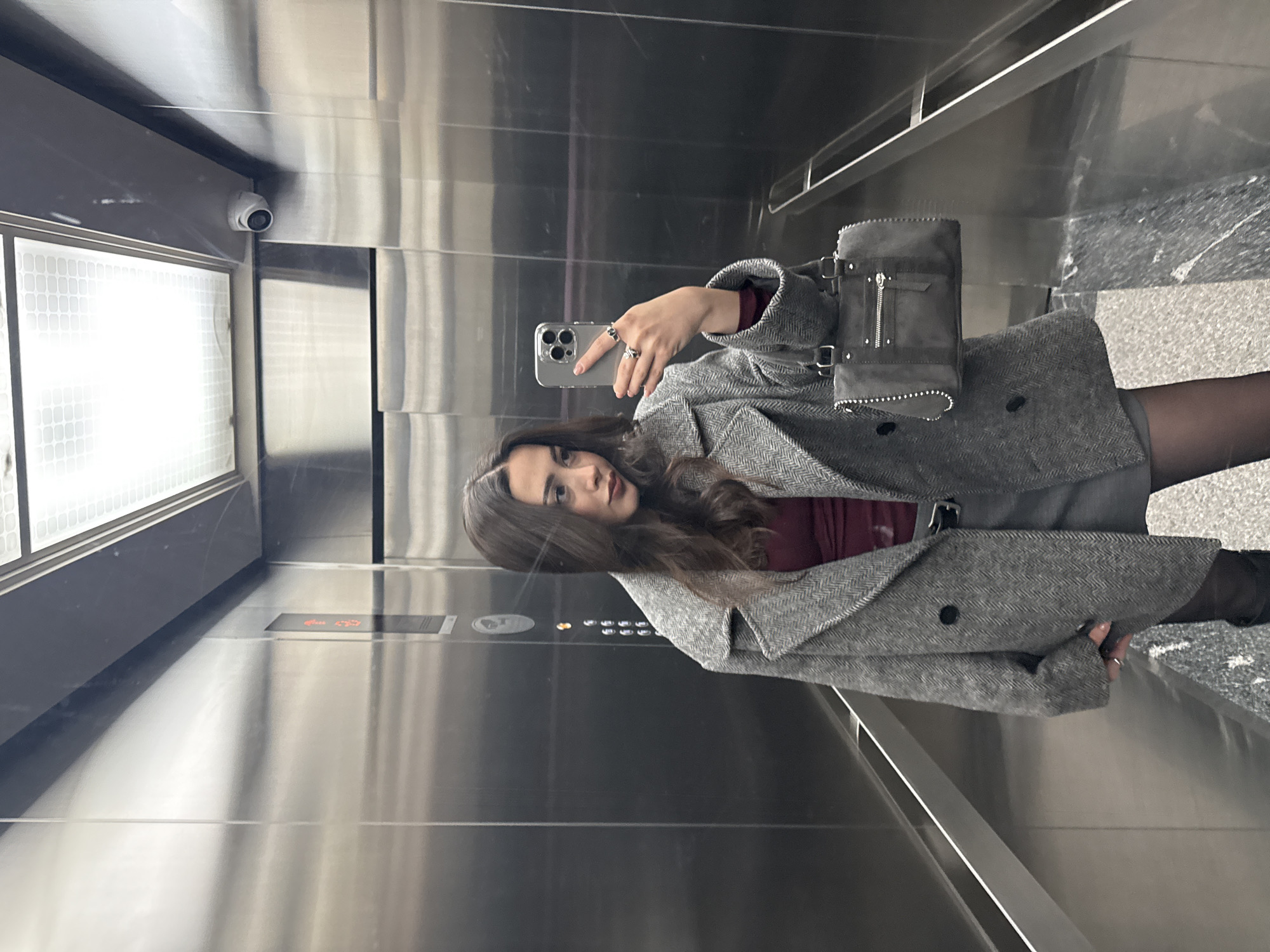 A woman is taking a selfie in an elevator. She is wearing a gray coat over a dark top and a gray skirt or dress. She is holding a phone in her right hand. Her hair is dark and wavy. The elevator has mirrored walls and a control panel. The elevator doors appear to be open, and the floor outside is visible. The location and city are not determinable from the provided image.