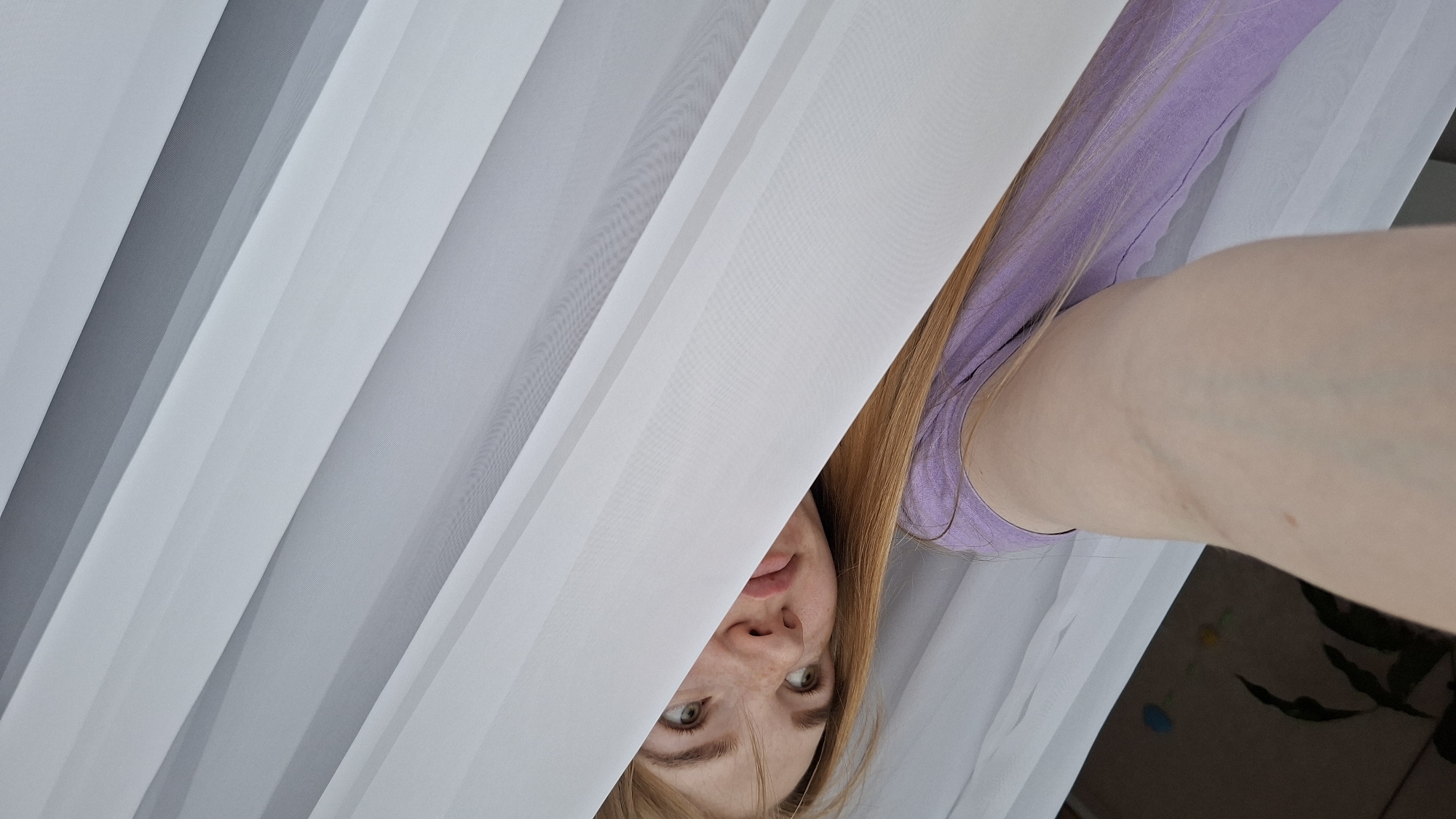 The image shows a young woman with long, reddish-blonde hair and green eyes peeking through white translucent curtains. She is wearing a light purple t-shirt. The camera angle is from her perspective, looking upwards and to the side, with a portion of her arm and shoulder visible in the foreground. The curtains create a sense of privacy or hiding. The background is out of focus but appears to be an indoor setting with some hints of furniture and plants. There is no identifiable information or context to place this image specifically in Chaikovsky, Russia.