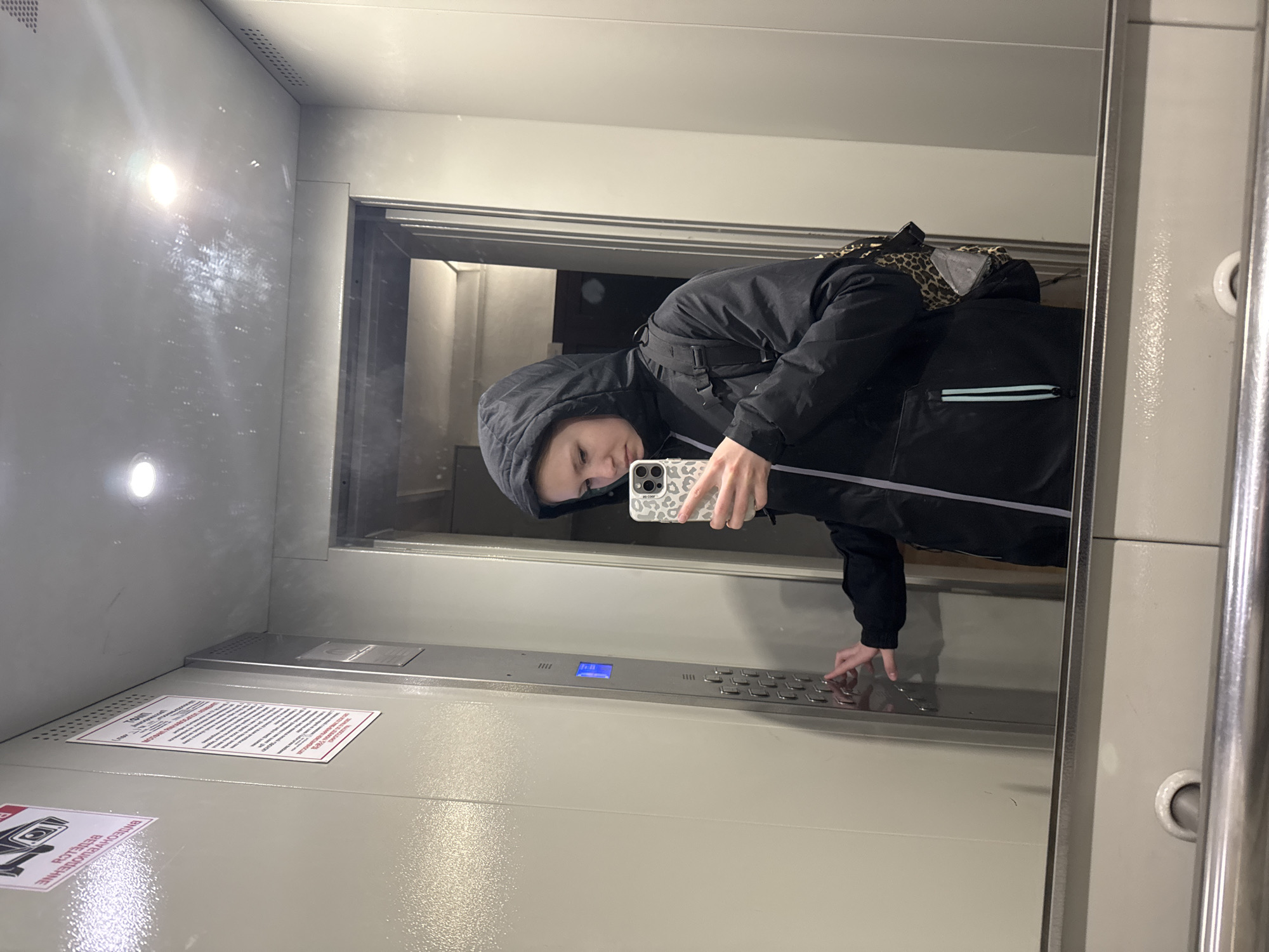 A person wearing a black hooded jacket is taking a selfie in an elevator. The elevator appears to be modern with silver buttons and a blue display panel. A sign with dense text is visible on the elevator wall. The location is presumed to be Chaikovsky, Russia. The person is pressing a button on the elevator panel.