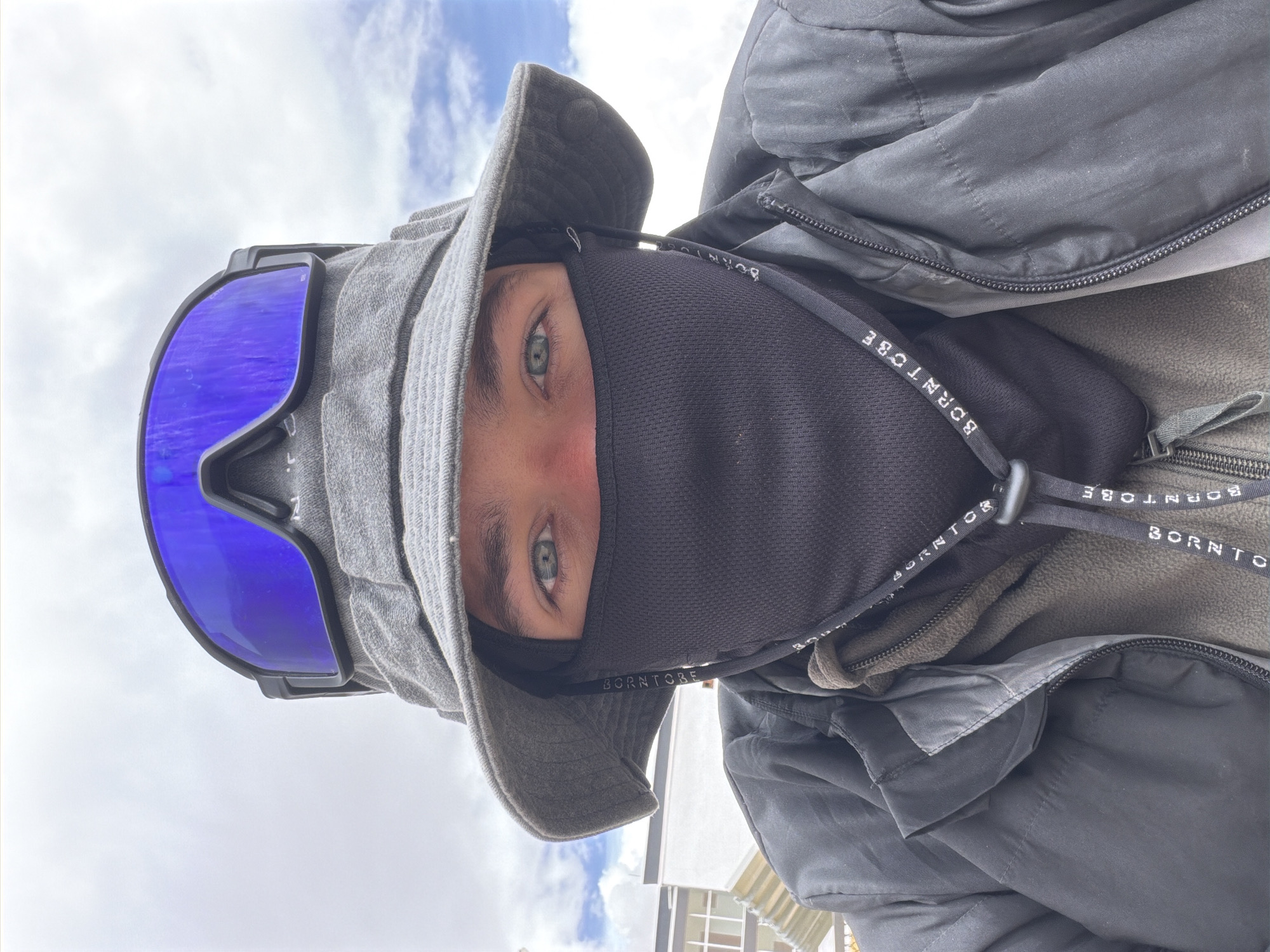 A close-up, selfie-style photo shows a person wearing a gray bucket hat and blue ski goggles. They are also wearing a black face mask that covers their nose and mouth, and a dark jacket over a green hoodie. The background suggests an outdoor setting with a cloudy sky and a building with windows. The image appears to have been taken in Karzok, India, possibly near the shore of Tso Moriri lake, given Karzok's location in the region.